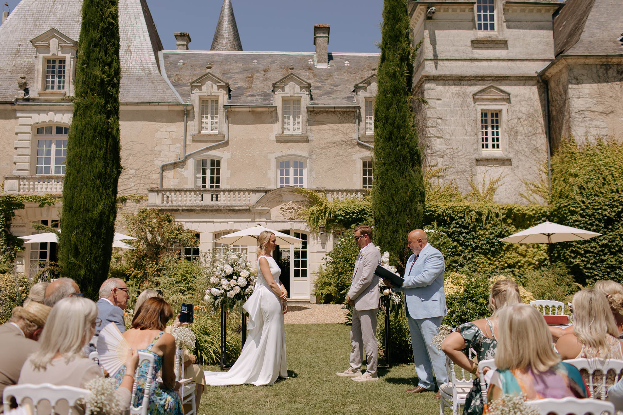 An outdoor wedding ceremony taking place on the lawn in front of a French château, with the couple standing at the altar facing an officiant dressed in a light blue suit who is reading from a black folder. The bride wears a sleek, fitted white gown with a short train, and the groom is dressed in a light beige suit. A tall black arch or stand decorated with white and cream florals — including what appear to be peonies or hydrangeas — marks the ceremony space between two tall cypress trees, with the château's cream limestone façade and arched doorways forming the backdrop. Approximately 20–30 seated guests in white Chiavari chairs are visible on either side of the aisle, dressed in summery colorful attire, with white parasols positioned to the sides. The composition is a wide shot taken from behind the seated guests, capturing both the couple and the full château façade. Potential venue feature image.