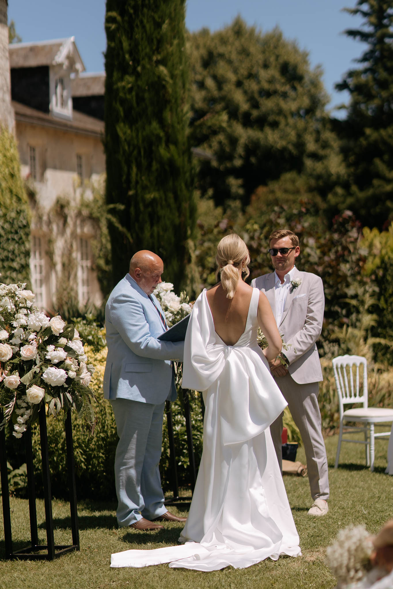 An outdoor wedding ceremony taking place on a lawn in front of a French château or manor house, with tall cypress trees visible in the background. The bride stands with her back to the camera wearing a white gown with a striking oversized bow detail at the back and a low open back, with a train extending across the lawn and her blonde hair in a low chignon; the officiant in a light blue suit reads from a folder to her left, while the groom stands facing her wearing a light grey suit with a white boutonniere and sunglasses. To the left of the ceremony, white roses and greenery are arranged on black geometric pedestal stands, contributing to a modern, minimal floral palette. The shot is a medium-wide portrait taken from behind the couple, with white Chiavari chairs visible to the right and a few guests partially in frame in the foreground.