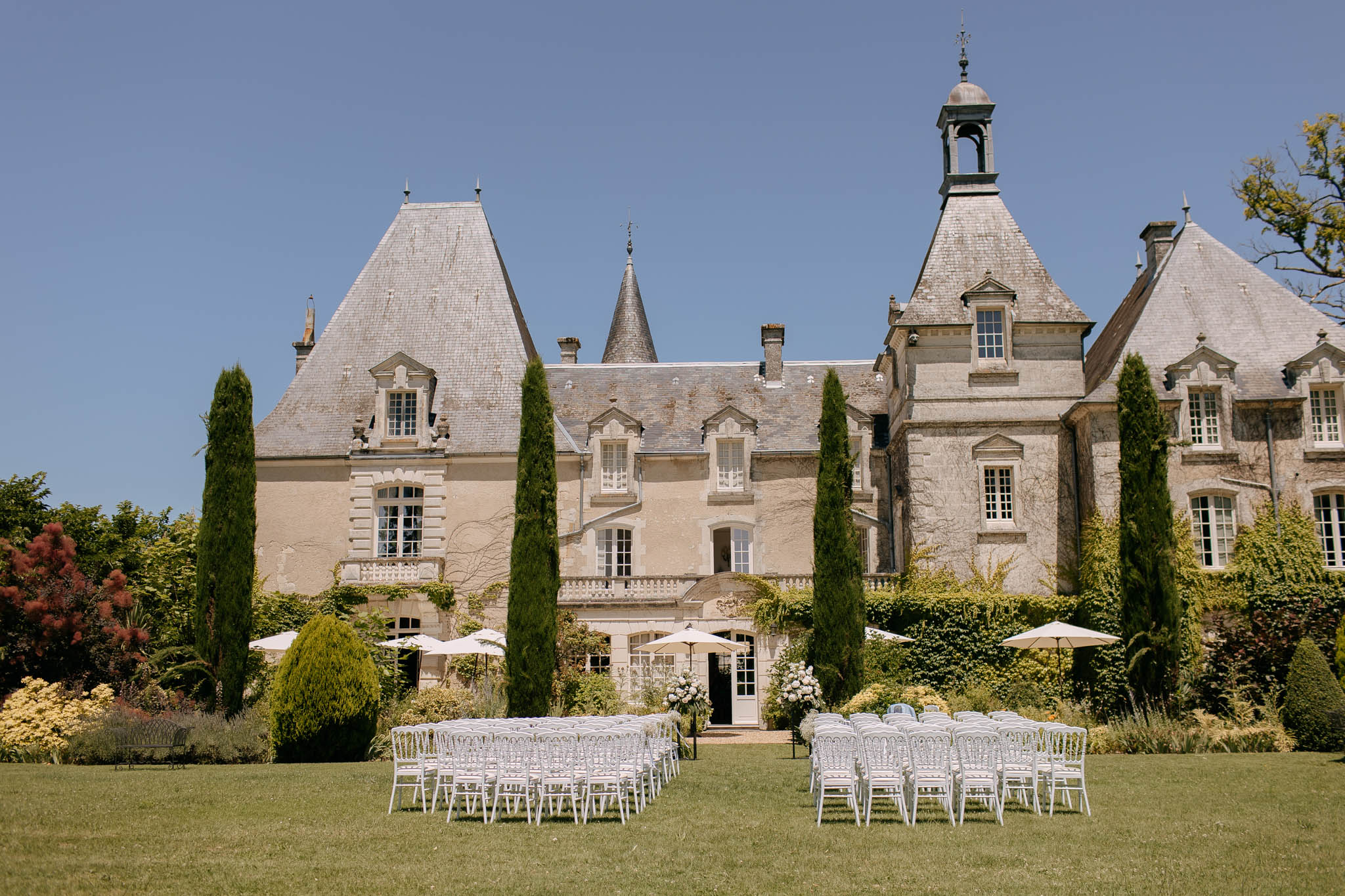 Pre-ceremony setup Napoleon chairs white hydrangea pedestals cream umbrellas before multi-turreted slate-roofed chateau