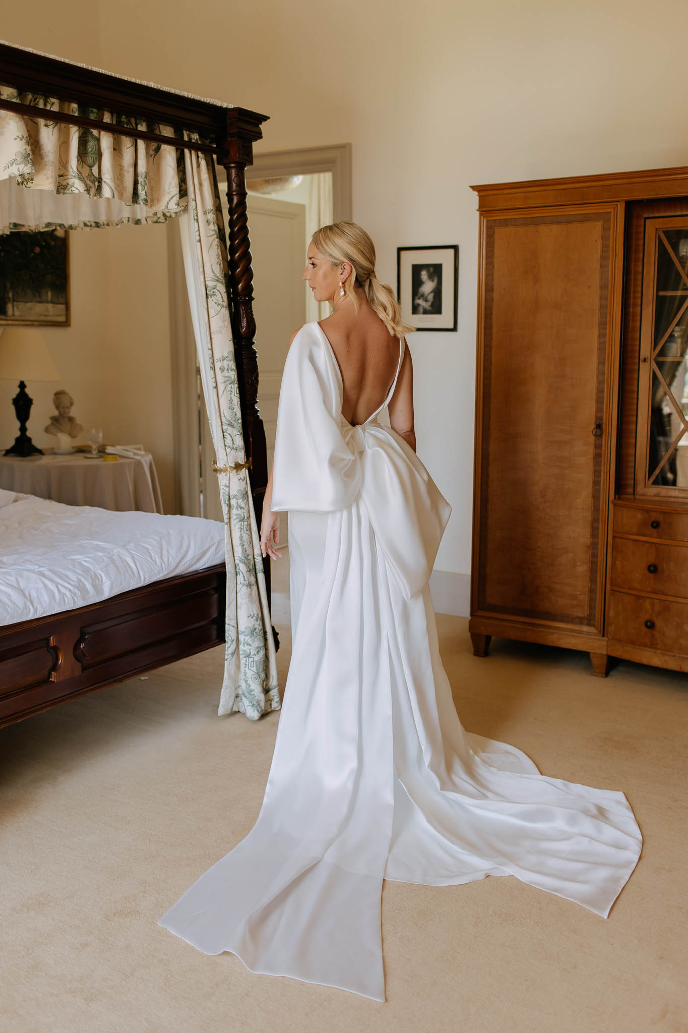 A getting-ready portrait of a bride standing alone in a bedroom, shot from behind at a three-quarter angle. She wears a white satin gown with a deep open back, oversized bow detail at the waist, long cape-like sleeves, and an extended train pooling on the floor. Her blonde hair is styled in a low ponytail, and she wears drop earrings. The room features a dark wood four-poster bed with green and white floral canopy curtains, a warm honey-toned wooden armoire, framed artwork on cream-colored walls, and a bust sculpture on a side table. The overall styling is modern and minimalist. Full-length portrait composition.