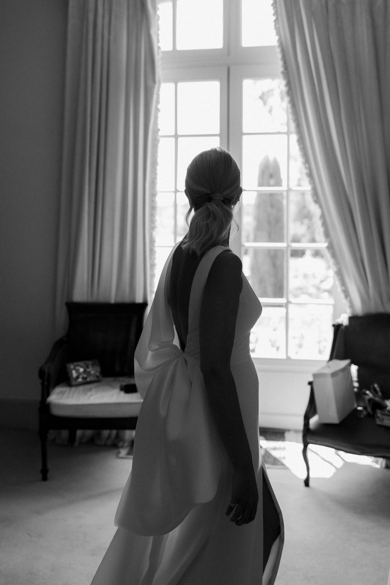 Black and white portrait of bride from behind showing open-back gown with bow detail near French window