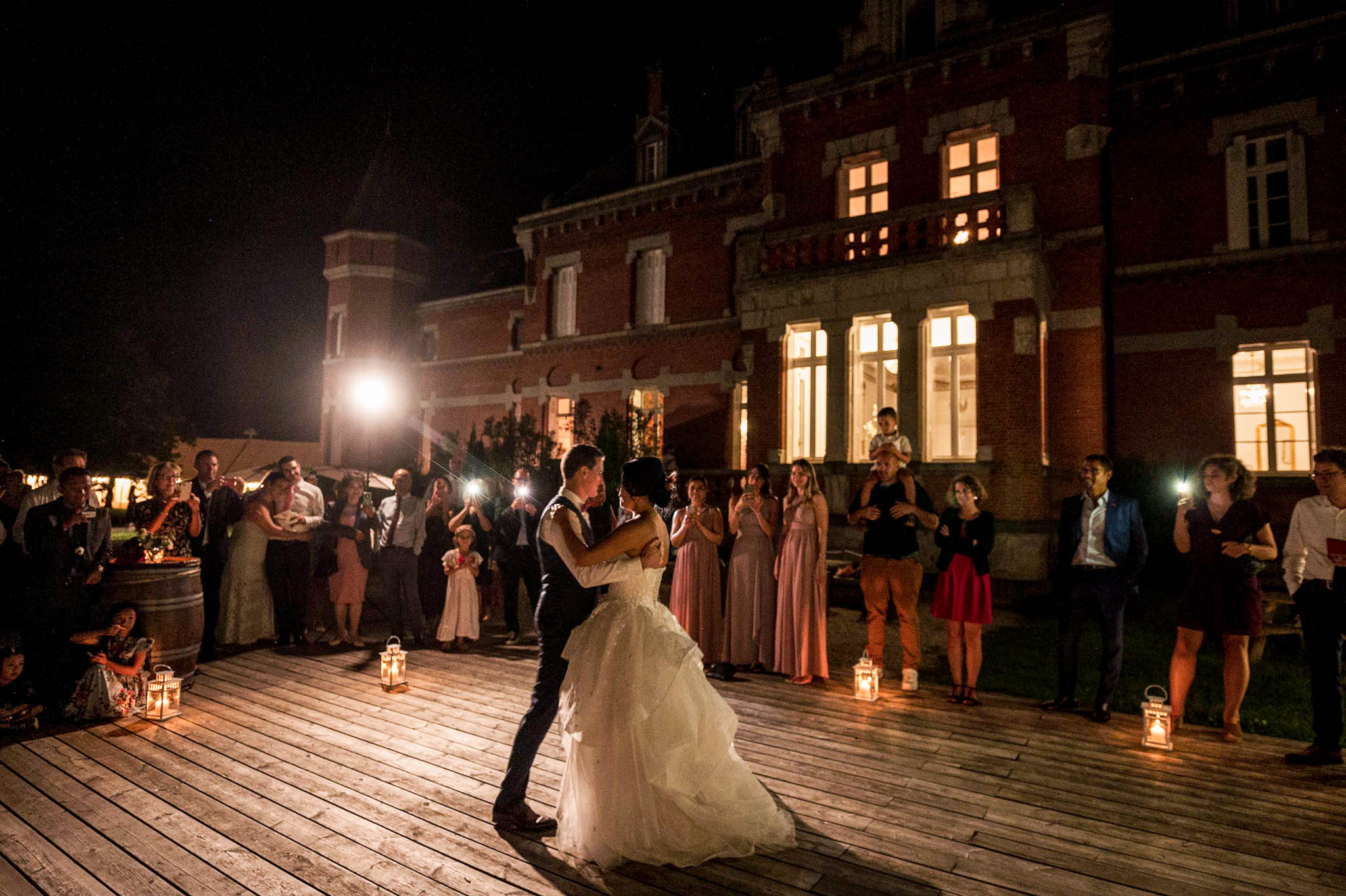 The couple shares their first dance outdoors at night on a wooden deck in front of a red brick château with illuminated windows. The bride wears a full-length white ballgown with a voluminous tulle skirt, and the groom is dressed in a dark suit. Approximately 30–40 guests form a circle around the couple, several holding up phone lights and cameras; bridesmaids in dusty rose floor-length dresses are visible among the crowd. White candle lanterns are placed around the perimeter of the dance floor, providing warm ambient lighting alongside the glow from the château's windows, giving the scene a classic, romantic atmosphere at a French château venue. The image is a wide shot taken at ground level, capturing both the dancing couple in the foreground and the lit façade of the château in the background.