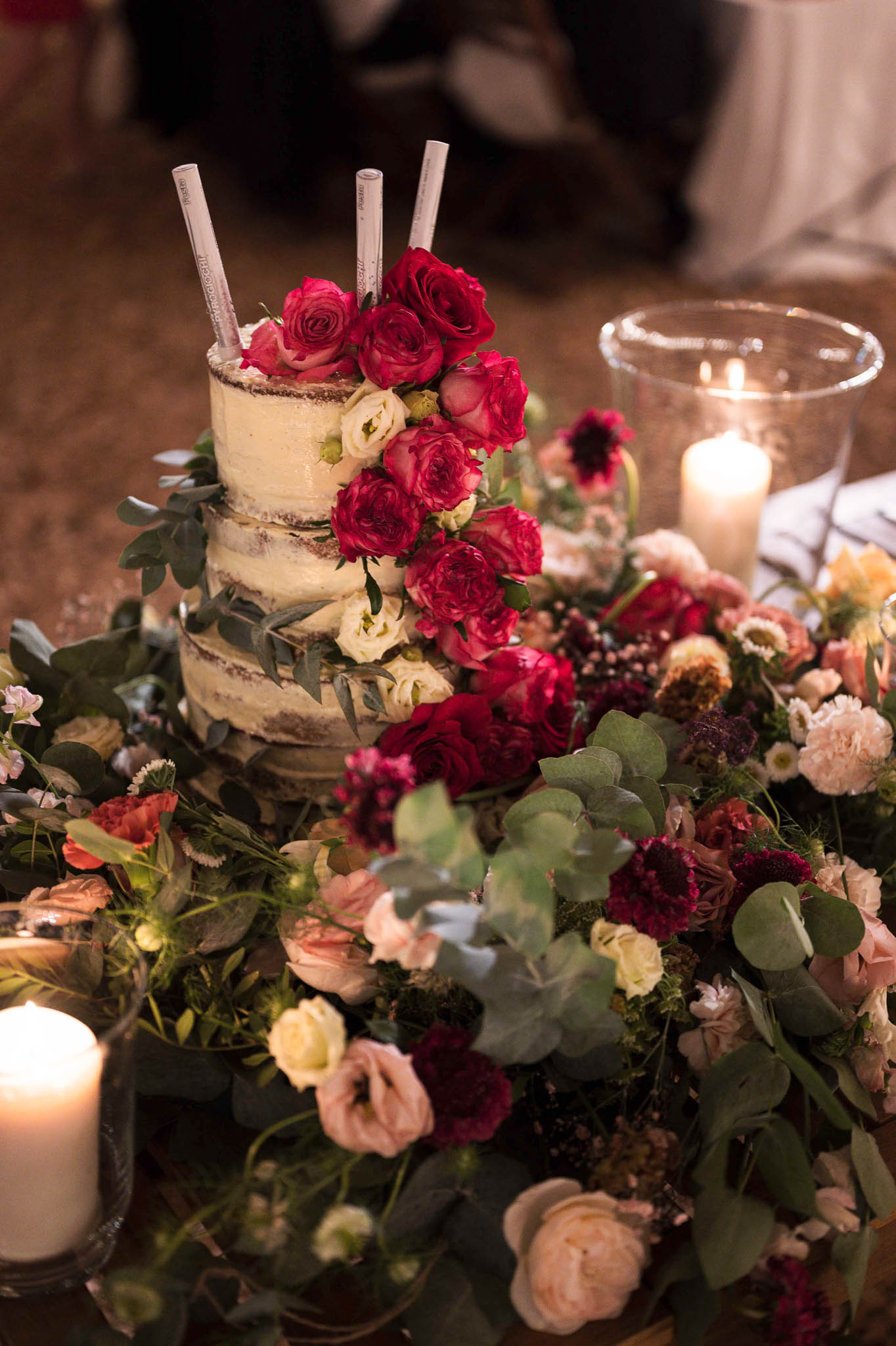 Close-up detail shot of a two-tier semi-naked wedding cake with a thin ivory buttercream finish, decorated with a cascading arrangement of deep red roses, cream ranunculus, and eucalyptus foliage. Three small labeled sparkler sticks are inserted into the top of the cake. The cake is surrounded by a lush floral table garland featuring deep burgundy carnations, blush garden roses, dusty pink ranunculus, cream spray flowers, and abundant eucalyptus and mixed greenery. Two pillar candles in glass hurricane holders flank the cake, casting warm ambient light across the display. The overall decor palette is rich jewel-toned with burgundy, deep red, blush, and cream against dark greenery, suggesting a romantic, garden-inspired reception styling. The background is softly blurred, with hints of guests and white table linens visible.