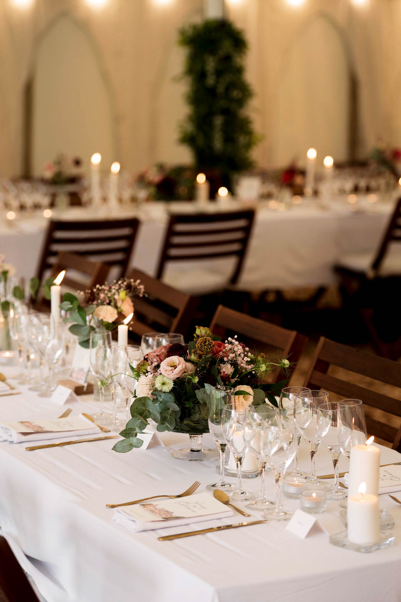Reception tablescape with gold cutlery, pillar candles, and low arrangements of blush roses and burgundy blooms