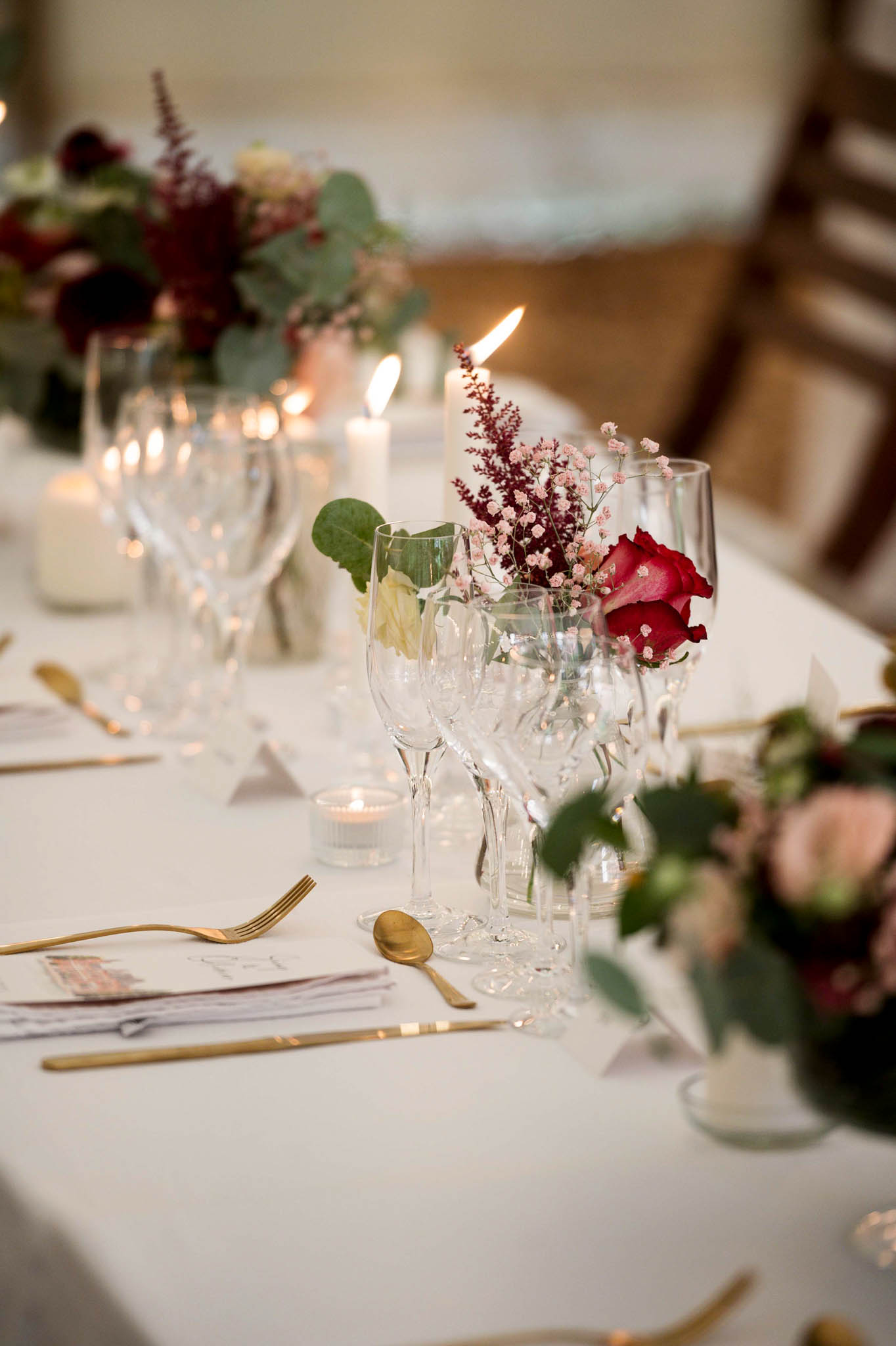 A close-up detail shot of a wedding reception table set with a white linen tablecloth, gold flatware, and multiple clear crystal wine and water glasses per place setting. Small bud vases hold deep red roses, blush pink gypsophila, burgundy astilbe, and eucalyptus foliage, while larger low centerpieces in the same burgundy, blush, and green palette are visible further along the table. Pillar candles and tea light votives provide warm candlelight throughout. Printed menus or place cards are visible beneath the gold cutlery. The overall decor palette is burgundy, blush, and gold with a classic, candlelit reception style.