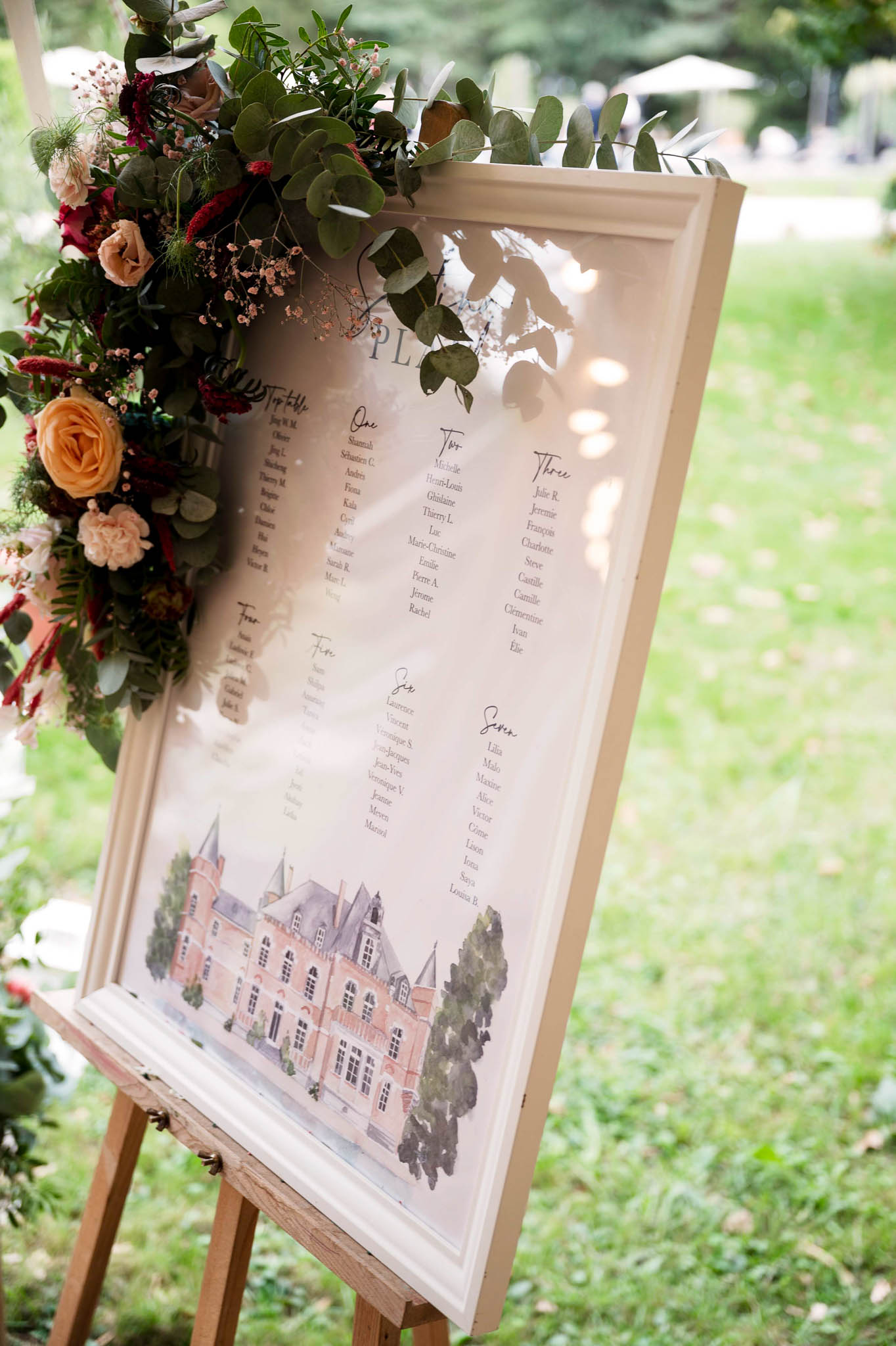 A close-up detail shot of a wedding seating chart displayed on a wooden easel, photographed outdoors in a garden setting. The framed white sign features a watercolor illustration of a French château at the bottom and lists guest names organized by table — including a top table and tables numbered one through seven — written in a calligraphy-style font. The upper left corner of the frame is adorned with a floral arrangement composed of burgundy blooms, peach garden roses, blush ranunculus, dried reddish amaranth, and trailing eucalyptus, creating a rich, romantic color palette of deep red and soft peachy tones. The decor style is classic with a romantic, slightly rustic feel, combining the custom venue illustration with lush organic florals.