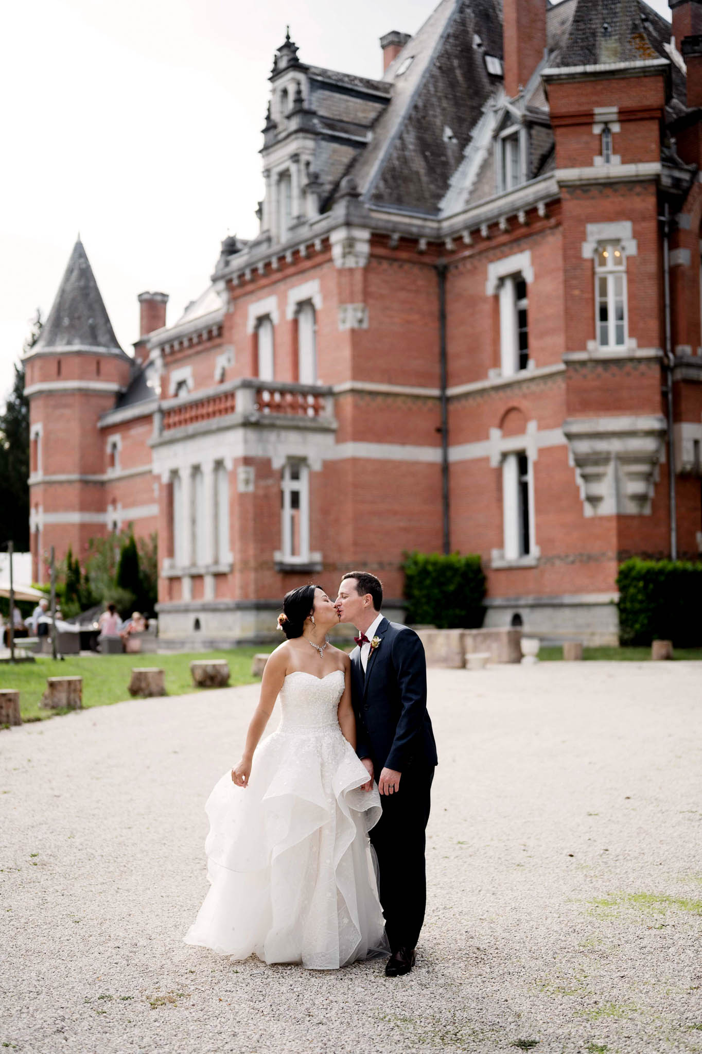 A couple portrait taken outdoors on the gravel forecourt of a French château, with the bride and groom kissing. The bride wears a strapless ivory ball gown with a layered, ruffled skirt and a jeweled necklace, with a floral hair accessory featuring a burgundy bloom. The groom wears a navy suit with a burgundy bow tie and a small boutonnière. In the soft-focus background, the château is a multi-story red brick building with slate-roofed turrets, white stone trim, and dormer windows in the French château style; a small group of guests is visible gathered to the left side of the building. The composition is a full-length portrait shot with the couple centered in the foreground and the château façade filling the background. Potential venue feature image.