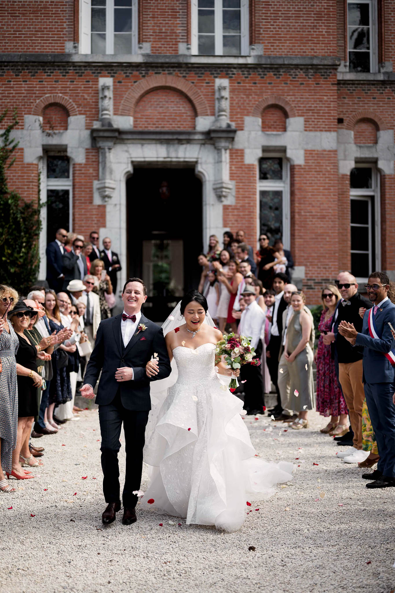 The bride and groom are exiting a red-brick château-style building through a guest-lined gravel pathway in an outdoor post-ceremony recessional. Guests — approximately 40–50 people — line both sides of the path, clapping and tossing red and ivory rose petals. The bride wears a strapless, full-ballgown dress with a layered ruffled tulle skirt and holds a bouquet of deep burgundy and blush blooms with greenery; she also wears a cathedral-length veil. The groom is dressed in a dark navy suit with a burgundy bow tie and a floral boutonnière. A man on the far right wears a French tricolor mayoral sash, indicating this follows an official civil ceremony. The shot is a wide, full-length portrait taken from ground level, with the red-brick building with stone arched detailing and decorative stonework visible in the background.