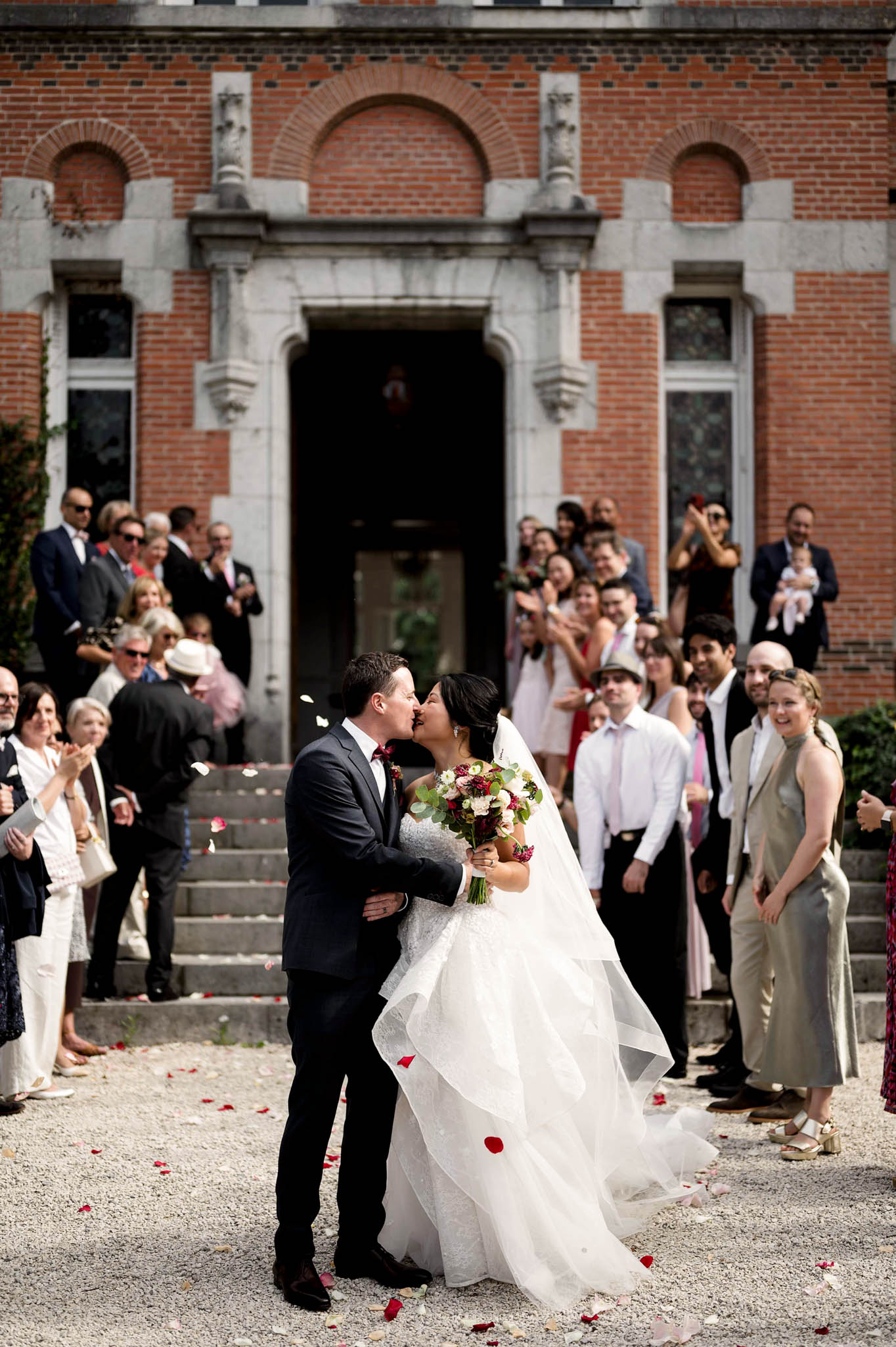 Bride and groom kissing during petal-strewn exit before red brick chateau with cheering guests