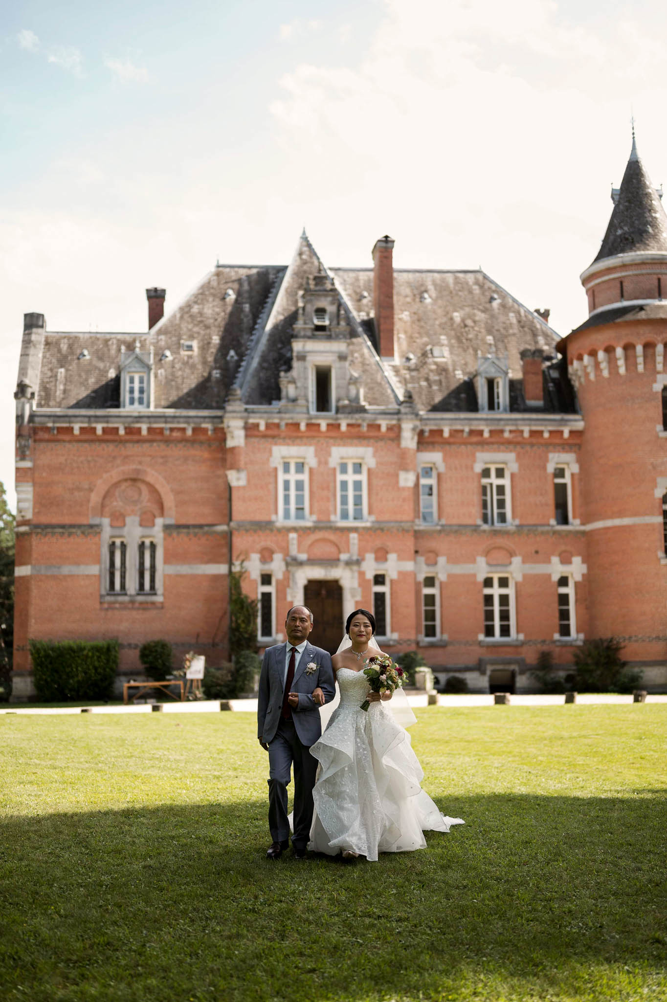 A bride is escorted across a lawn by a male companion — likely her father — in front of a large French château built in red brick with white stone detailing, slate mansard roofs, and a rounded corner turret. The bride wears a strapless white ball gown with a tiered, ruffled skirt featuring a subtle dotted texture, and carries a loose bouquet with burgundy and mauve tones. Her escort wears a blue-grey suit with a burgundy tie and a boutonnière. A wooden easel with a sign is visible near the château entrance in the background. The composition is a wide, full-length portrait shot with the château facade filling the upper two-thirds of the frame. Potential venue feature image.