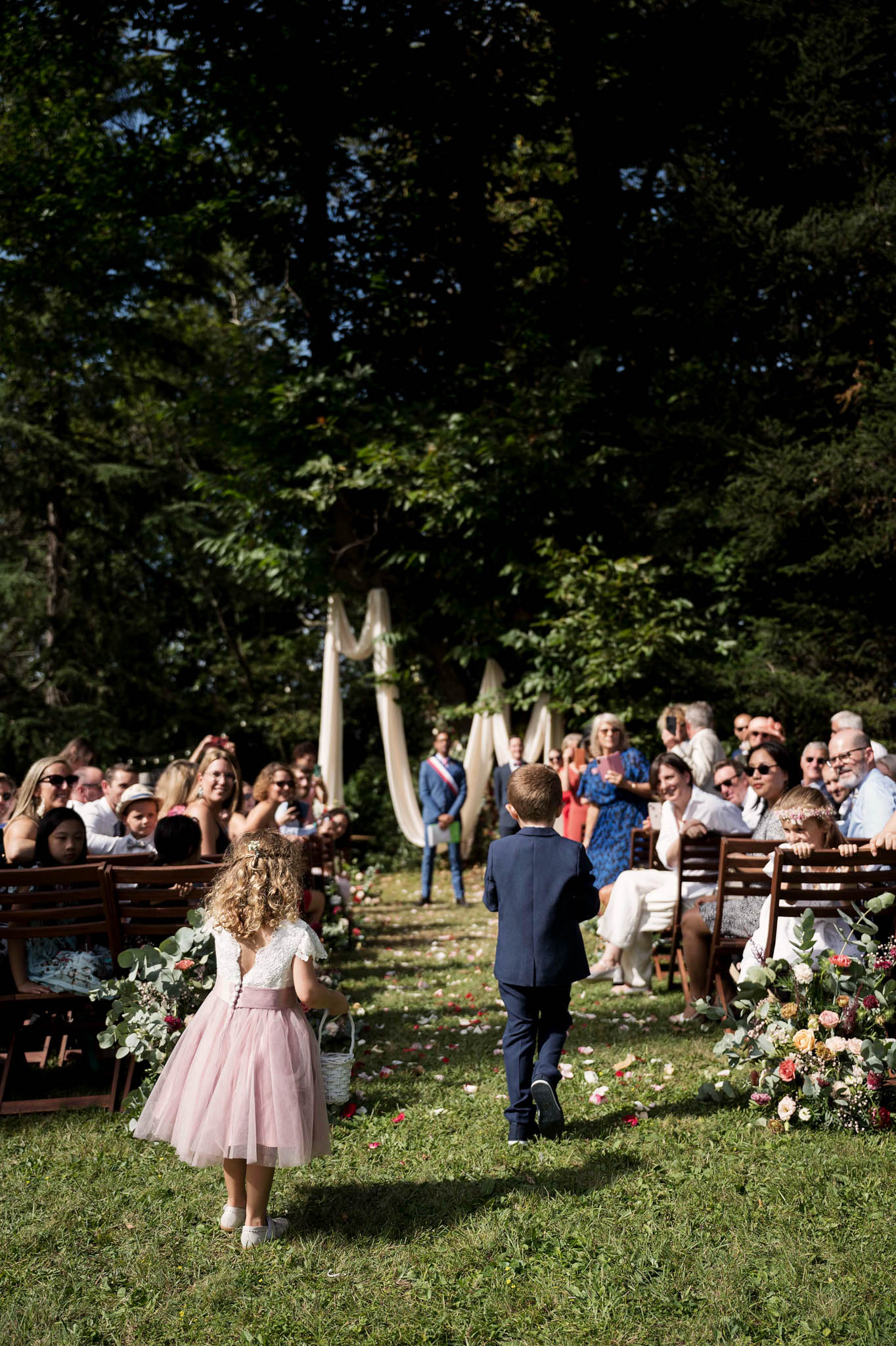An outdoor wedding ceremony is underway in a wooded garden setting, with a flower girl and ring bearer walking down a grass aisle toward the altar. The flower girl wears a white lace top with a blush pink tulle skirt and carries a small white basket, while the boy is dressed in a navy suit. The aisle is lined with lush floral arrangements featuring peach, blush, and burgundy roses with eucalyptus, and rose petals are scattered across the grass. Approximately 40–50 seated guests on dark wooden chairs flank both sides of the aisle, and a cream/ivory draped fabric arch is visible at the far end near the officiant. The shot is a wide, medium portrait taken from behind the two children, capturing the full aisle scene in natural daylight.
