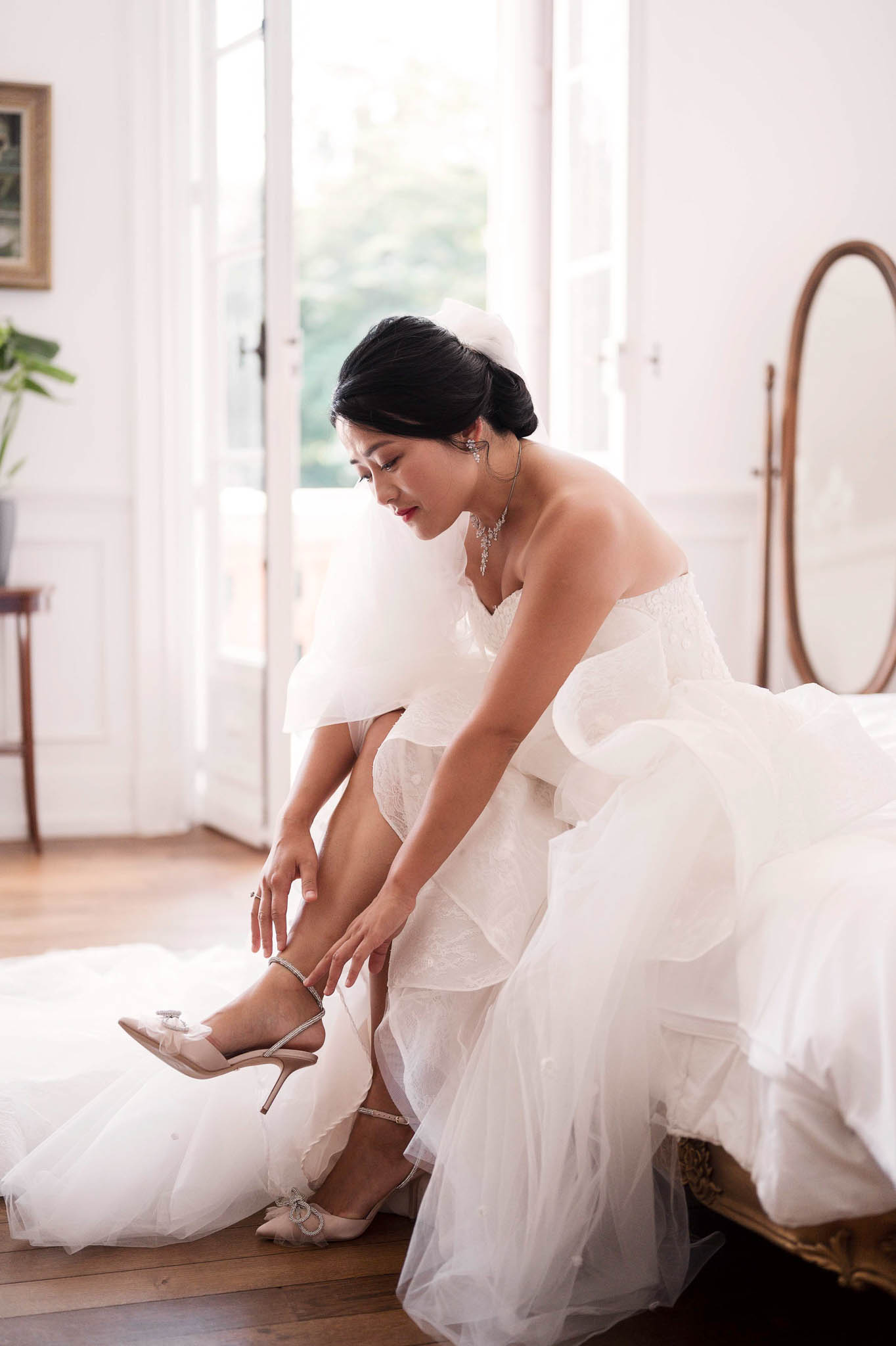 A getting-ready moment captured indoors, showing a bride seated on the edge of a gilt-framed bed fastening the ankle strap of a nude satin stiletto heel with a small bow detail. She wears a strapless white gown with layered ruffle organza detailing over a lace fitted underlayer, paired with a crystal chandelier necklace, drop earrings, and a white floral hair accessory at the nape of her low chignon. The room features white panelled walls, tall French doors letting in soft natural light, wide-plank hardwood floors, and an oval wood-framed standing mirror in the background. The shot is a medium portrait taken at a slight angle, with the bride in three-quarter profile looking downward toward her shoes.