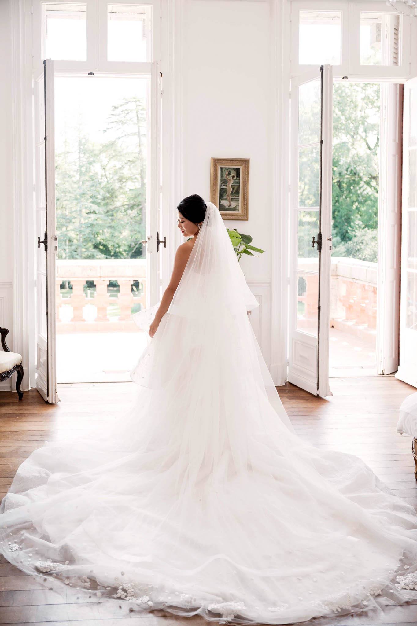 Bride from behind in strapless ballgown with floral applique train and cathedral veil, French doors open to terrace