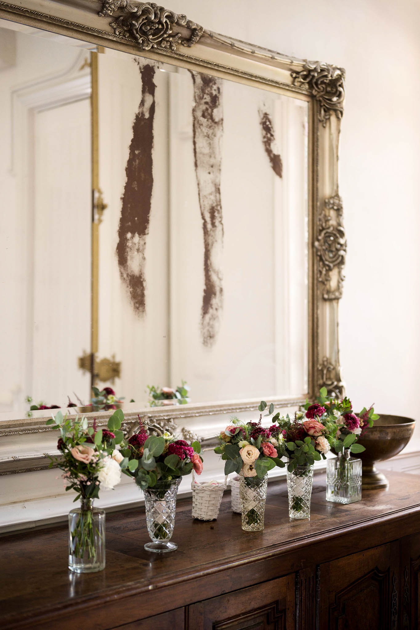 Blush roses and burgundy scabiosa in crystal vases on dark sideboard before aged gilt-framed mirror