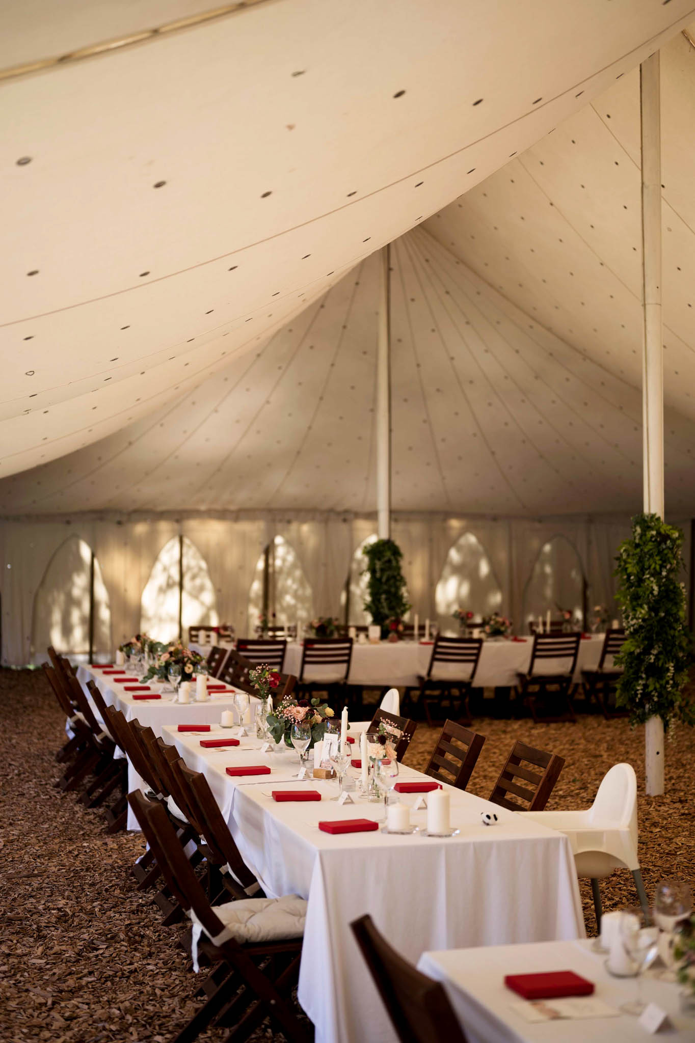 Sailcloth tent reception setup with long banquet tables, red napkins, blush floral centerpieces, and greenery on poles