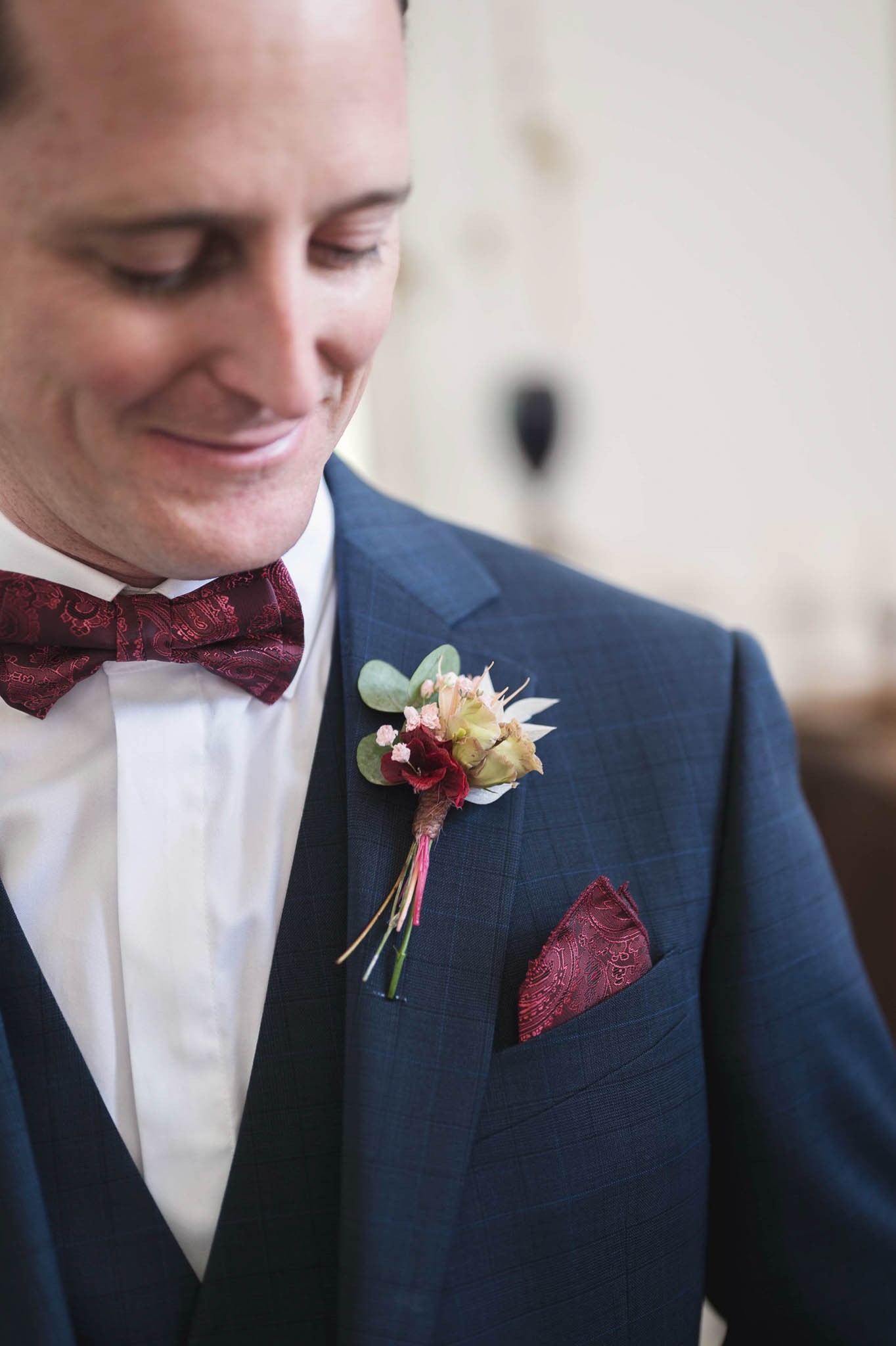 Close-up portrait of a groom dressed in a navy blue three-piece suit with a white dress shirt, a burgundy paisley bow tie, and a matching burgundy paisley pocket square. He is looking downward with a slight smile. His boutonniere features a small burgundy flower, a green rose bud, eucalyptus leaves, blush micro flowers, and dried grass stems. The background is softly blurred and appears to be a light-toned interior wall.