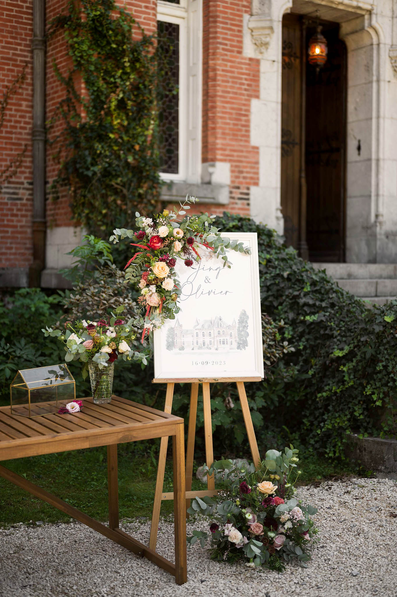 Wedding welcome sign on wooden easel with calligraphy names and date, flanked by blush and burgundy floral arrangements ou...