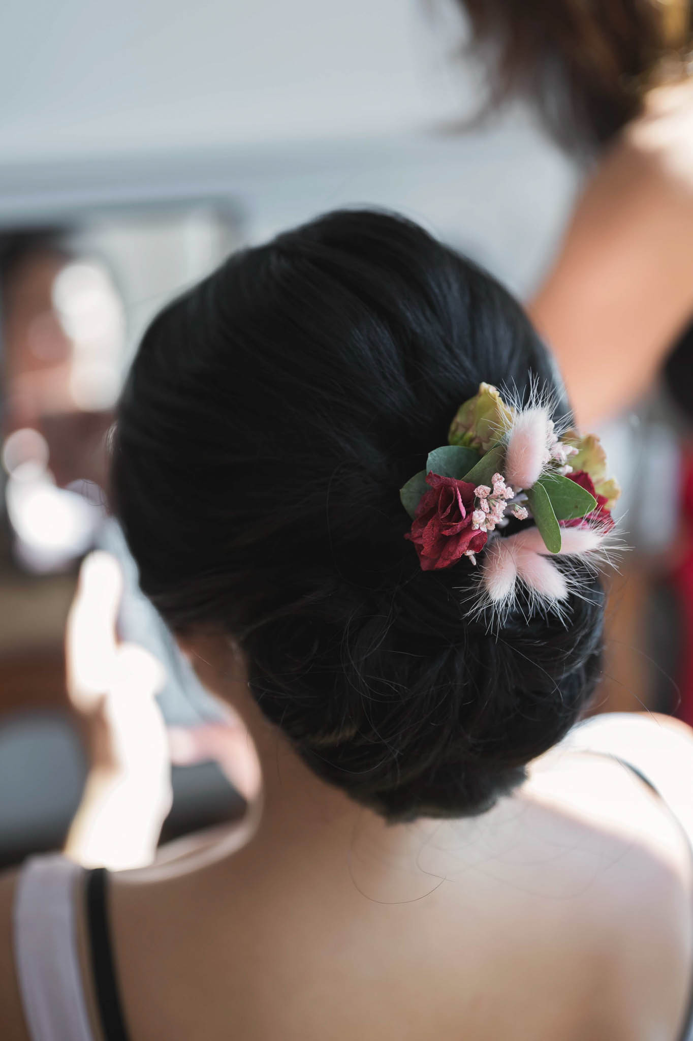 Close-up detail shot of a bride's hair styling during a getting-ready moment, taken indoors with natural light. The subject has dark hair styled into a low chignon, adorned with a floral hair piece featuring deep burgundy miniature roses, blush pink bunny tail grass (lagurus), small pink wax flowers, and eucalyptus leaves. The floral accessory combines a rich burgundy and blush pink color palette with soft, fluffy textures from the dried grasses. A second person is partially visible in the background, slightly out of focus, suggesting a bridal preparation setting.