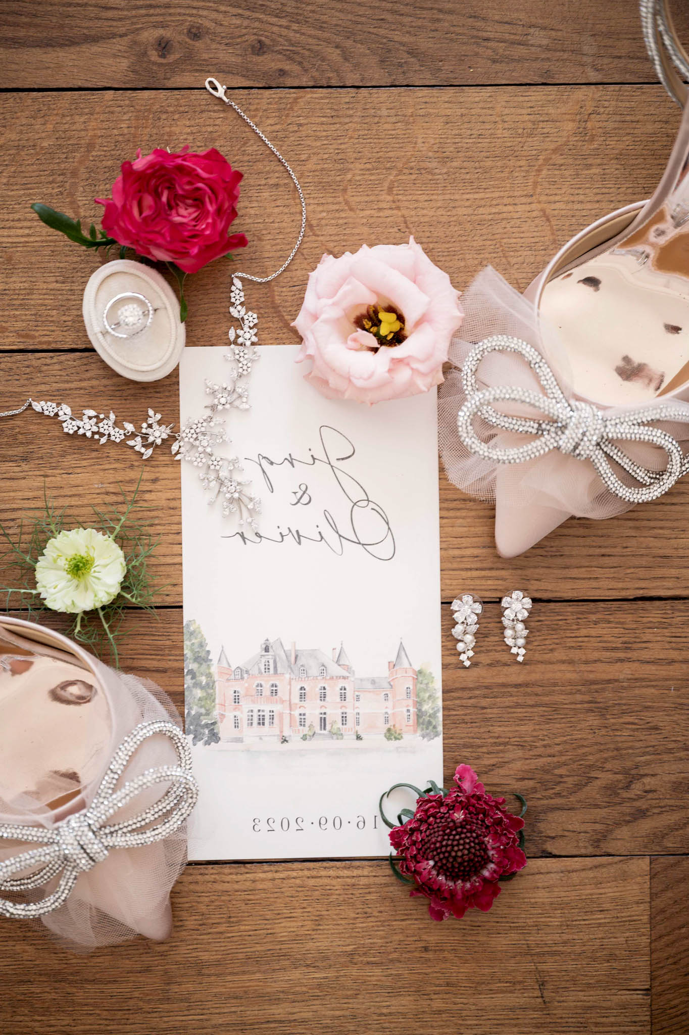 A flat lay detail shot arranged on a warm oak wood surface, featuring a custom wedding invitation card dated 16.09.2023 with calligraphy script names and a watercolor illustration of a pink château at the bottom. Surrounding the invitation are two pairs of blush pink heeled shoes with pearl-trimmed tulle bow detailing, a delicate silver floral-motif hair vine, a pair of small crystal drop earrings, a diamond-style ring displayed in a beige velvet ring box, a pearl strand necklace, a hot pink garden rose, a blush pink lisianthus bloom, a cream-toned ranunculus with green center, and a deep crimson sunflower-style bloom. The overall styling palette combines blush pink, deep magenta, and silver with a classic French château aesthetic.
