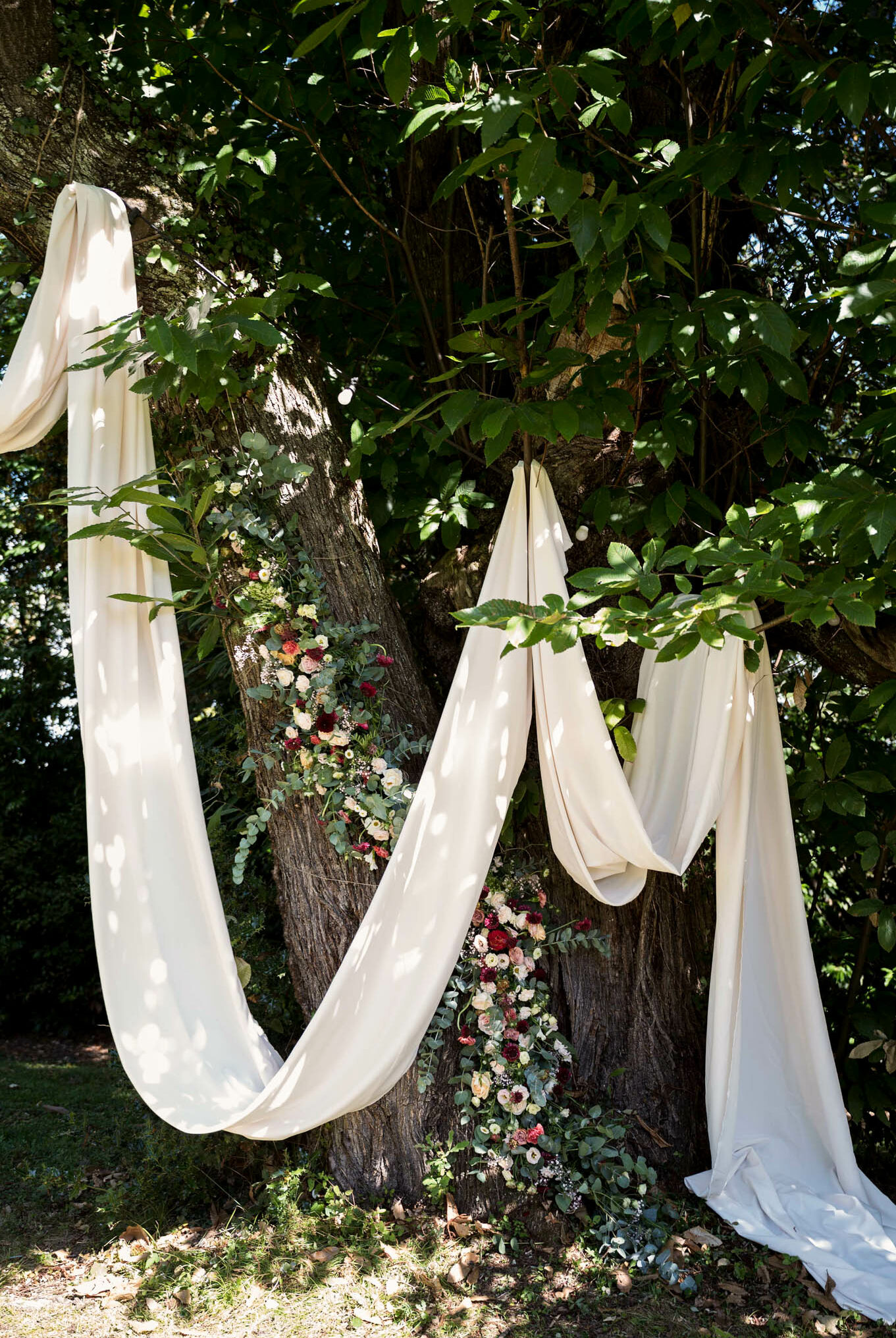 A close-up detail shot of an outdoor ceremony altar setup using large mature trees as a natural backdrop. Long panels of cream-ivory fabric are draped and swooped between the tree trunks, pooling slightly on the ground. Floral arrangements are placed at the base of the tree trunks, featuring deep burgundy, blush pink, dusty rose, and ivory blooms — likely dahlias, ranunculus, and garden roses — mixed with eucalyptus and trailing greenery. The overall styling is romantic-boho with a rich, jewel-toned floral palette contrasting against the soft cream drapery. Dappled sunlight filters through the tree canopy, casting light spots across the fabric.