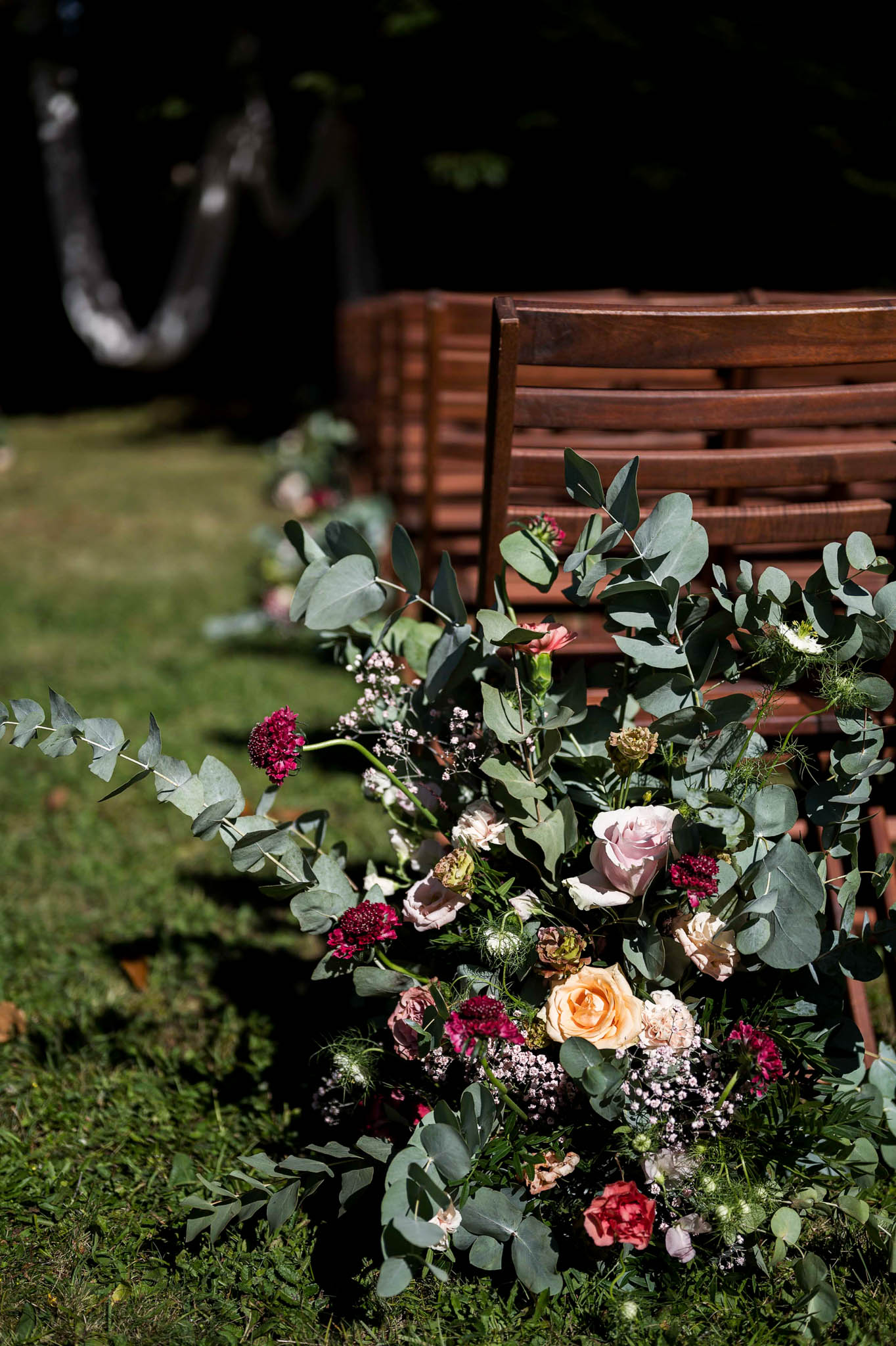 Ground-level aisle floral arrangement with eucalyptus, blush and peach roses, and burgundy blooms on ceremony lawn
