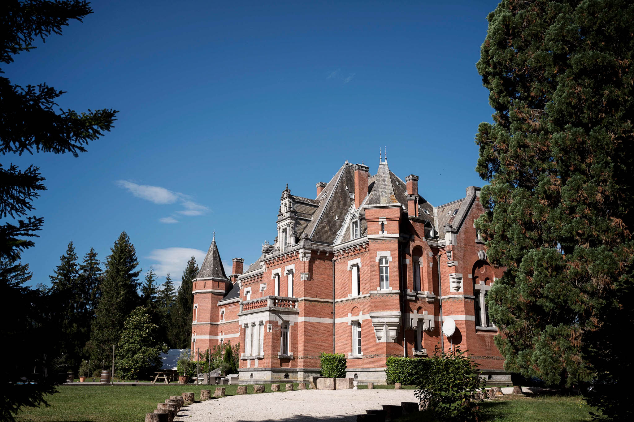 An exterior wide shot of a French château built in red brick with white stone detailing, featuring multiple steeply pitched slate roofs, pointed turrets, dormer windows, and ornate architectural trim typical of late 19th-century French château style. The building is photographed from the grounds, showing a gravel forecourt, a manicured lawn with tree stumps, and a wooden barrel visible to the left near what appears to be an outbuilding. No people are present in the frame. Potential venue feature image.