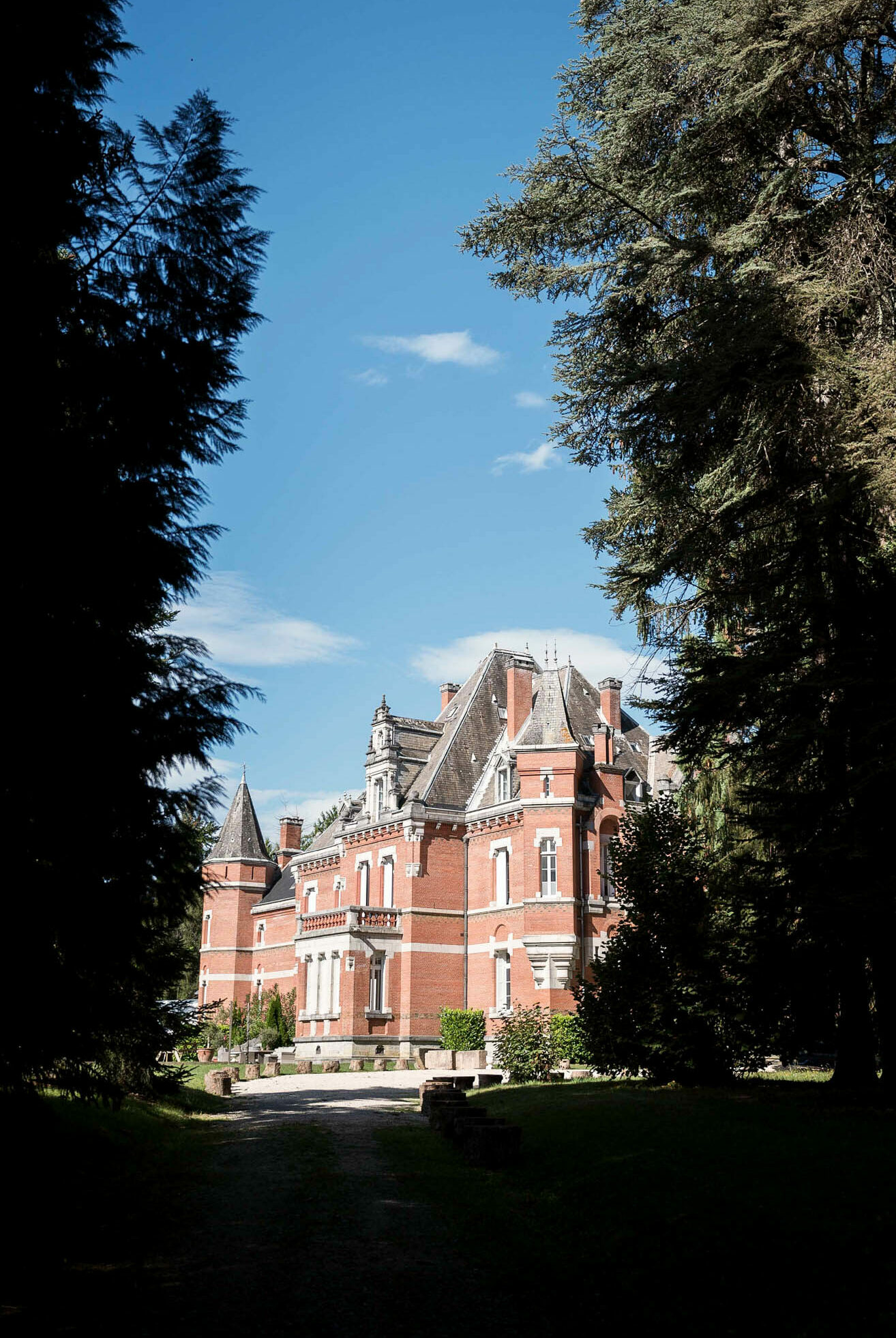 An exterior wide shot of a French château viewed from the grounds along a gravel driveway approach. The building is a large 19th-century-style manor constructed in red brick with white stone detailing, featuring steep slate mansard roofs, pointed corner towers, dormer windows, decorative cornices, and a balustraded upper terrace. The façade is multi-storey with arched and rectangular windows framed in white stone. The shot is framed by tall cedar trees on both sides, with the château centered in the middle distance. No people are visible in the image. Potential venue feature image.