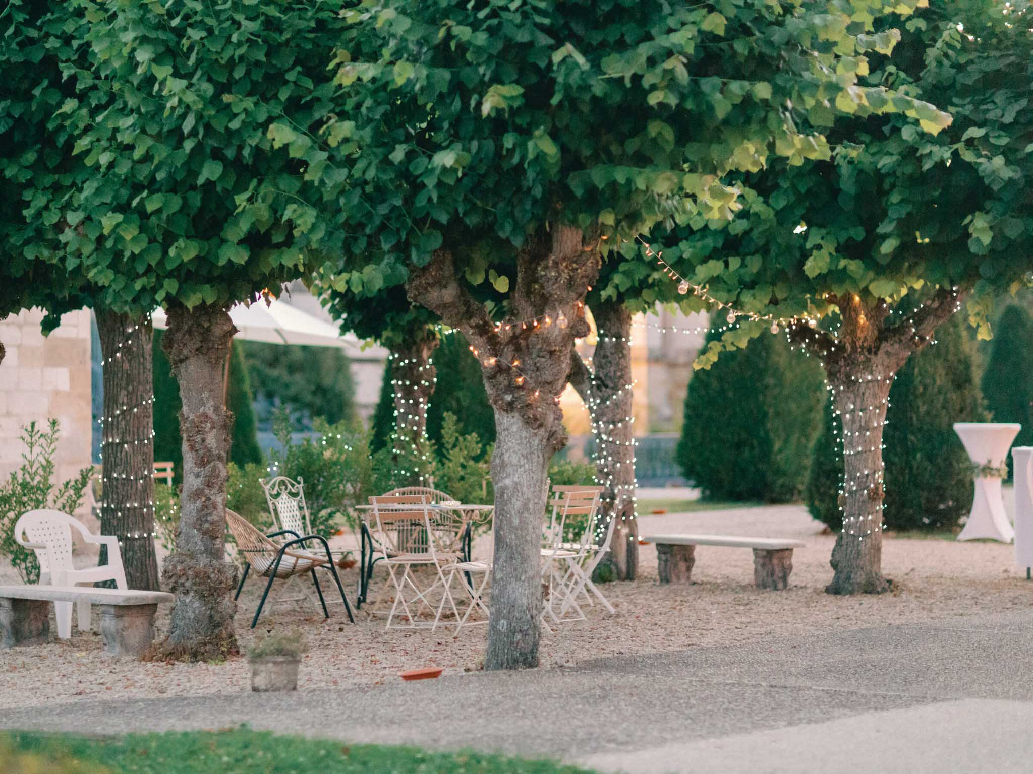 An outdoor cocktail hour or lounge area set up in a formal garden, photographed as a wide environmental shot at dusk. Several mature trees with broad canopies have warm white fairy lights wrapped around their trunks and branches, providing ambient lighting as natural light fades. Beneath the trees, a mix of white metal folding chairs, a rattan armchair, and a small bistro-style table are arranged informally on a gravel surface. A white plastic chair and stone bench are visible to the left, and a white cocktail-height poseur table with a linen cover is positioned to the right background. Trimmed conical topiary shrubs line the garden path, and a large white parasol is partially visible in the background alongside stone architectural elements. The overall styling is relaxed and garden-party inspired, mixing casual and ornamental furnishings. Potential venue feature image.