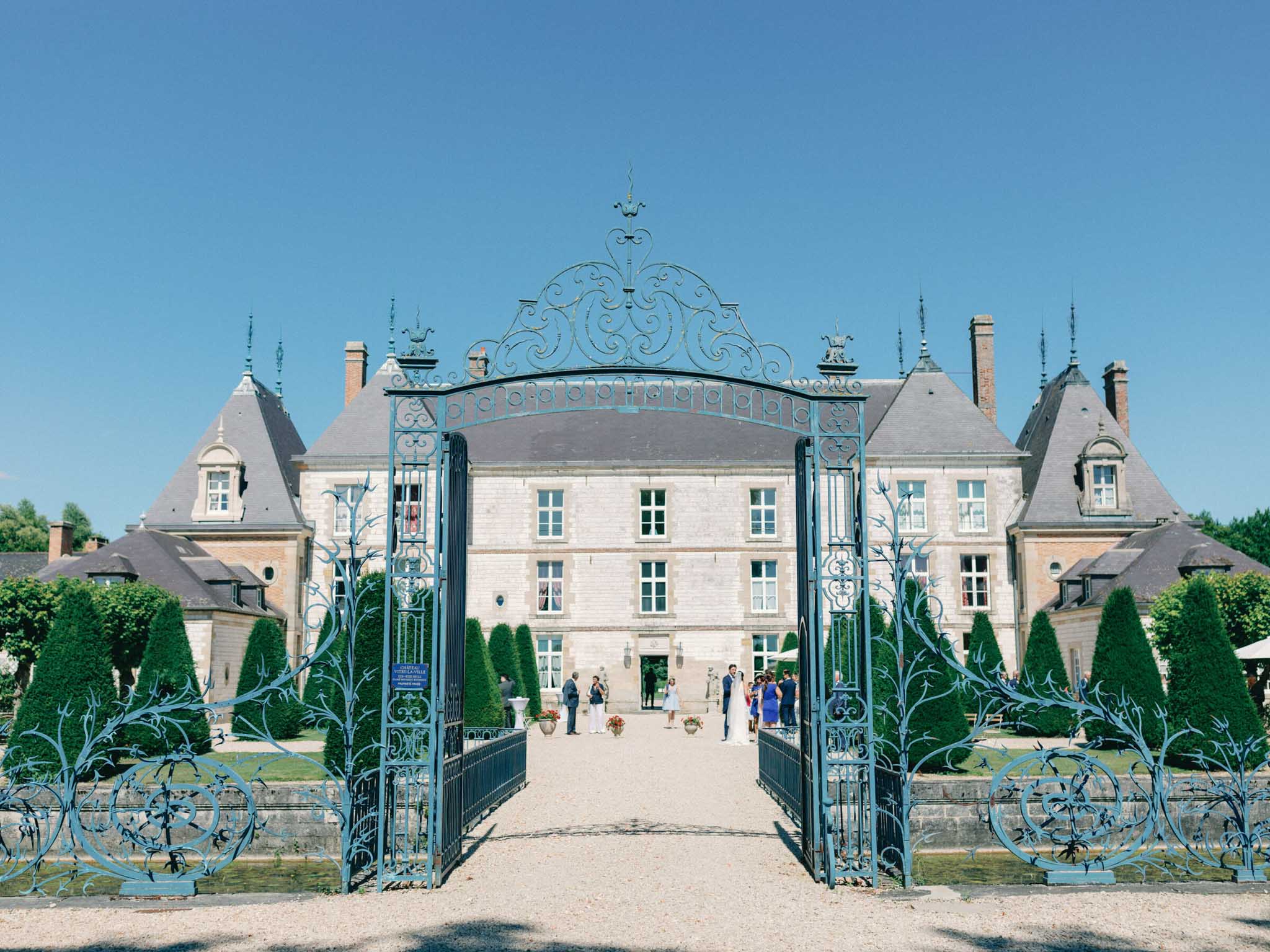 Classical French chateau viewed through ornate blue wrought iron gates with wedding party gathered at the entrance