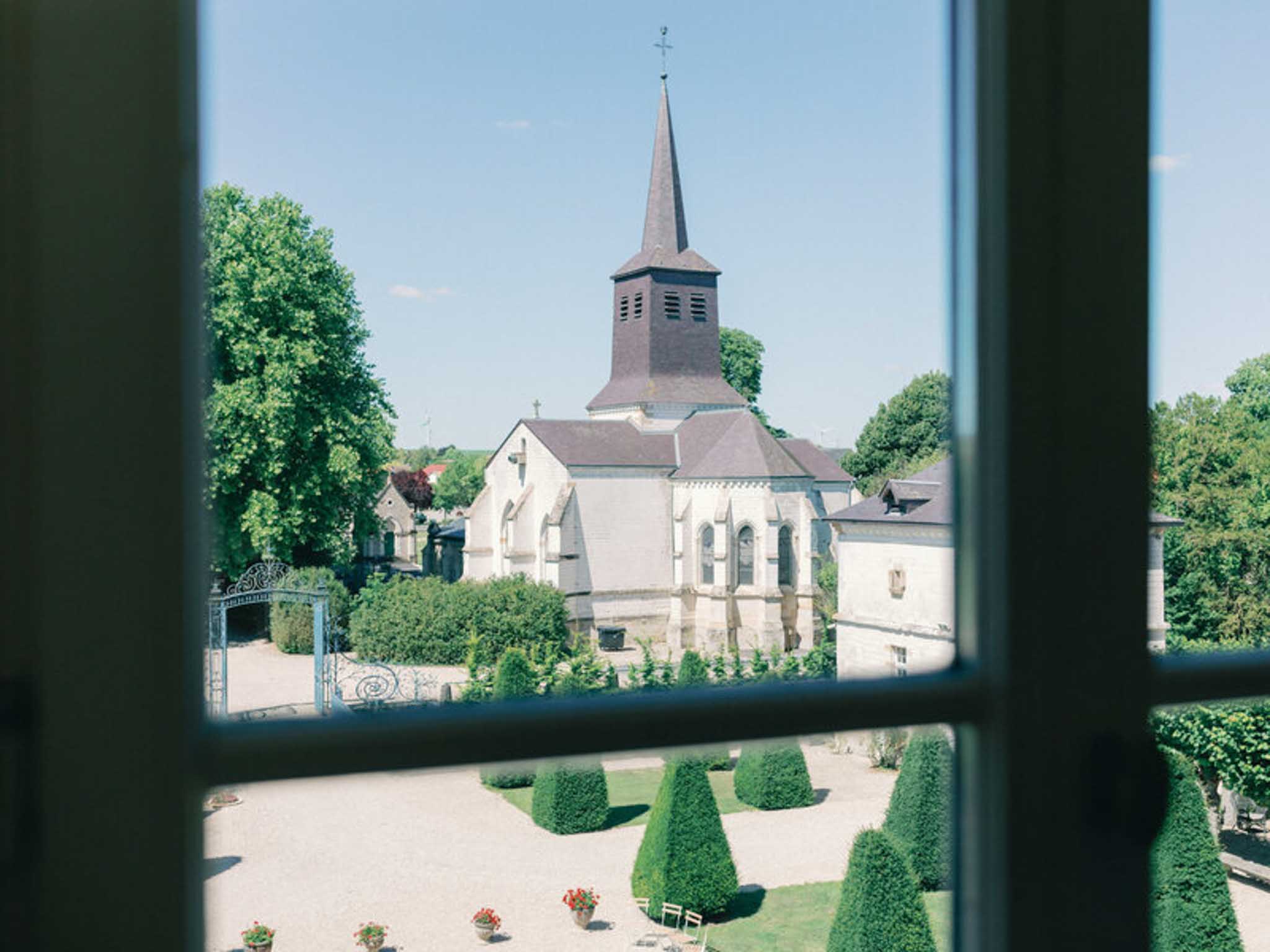 Village church spire viewed through multi-paned window with conical topiary and gravel terrace below