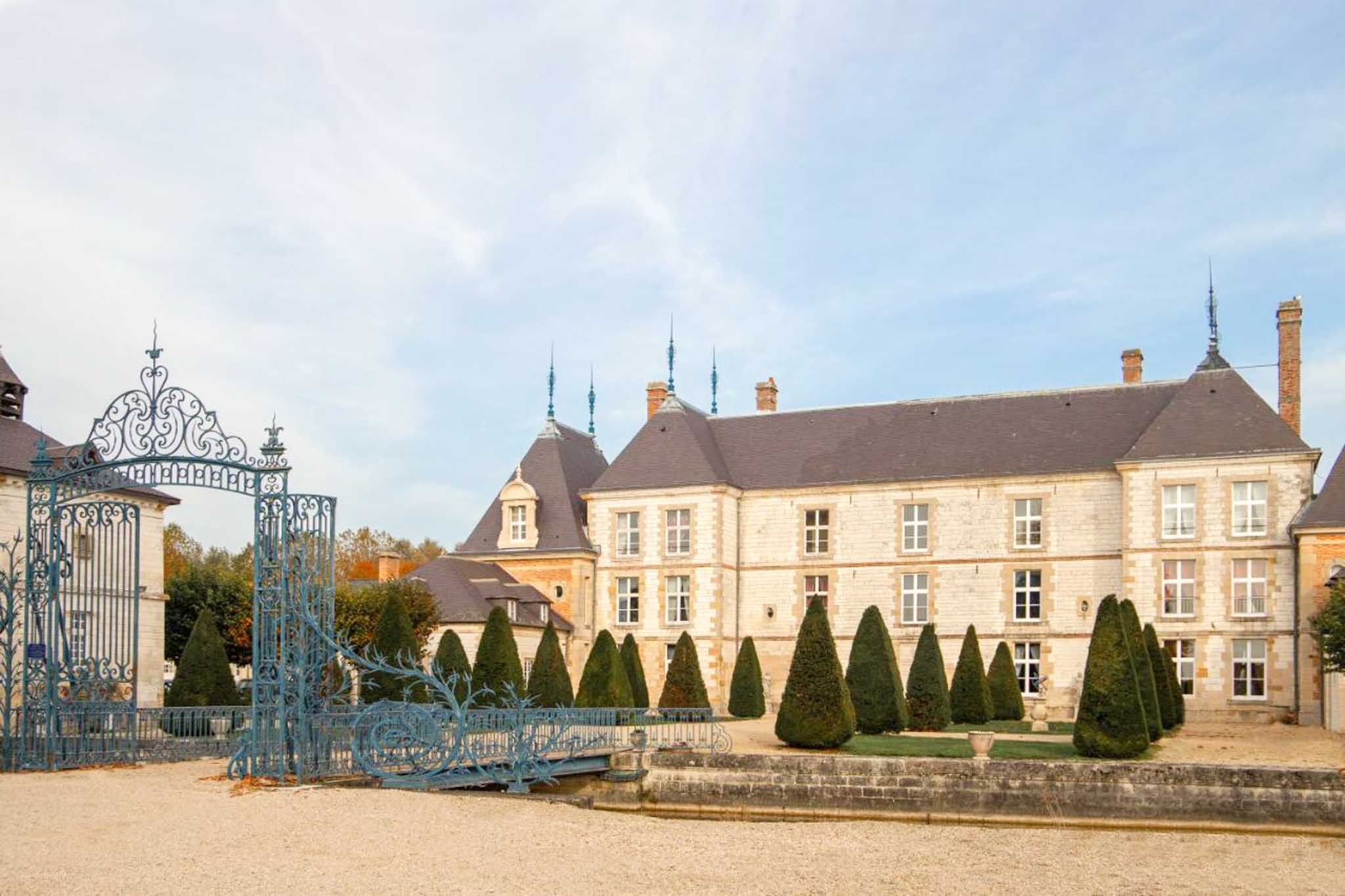 Wide exterior shot of a French château featuring a cream limestone facade with slate mansard roofs topped by decorative blue metal spires. The foreground features an ornate wrought-iron gate painted in teal-blue with scrollwork detailing, opening onto a gravel courtyard. A formal garden runs along the front of the building with a row of precisely clipped conical topiary shrubs. The building spans two to three stories with symmetrically placed white-framed windows across multiple wings. No people are visible in the image. Potential venue feature image.