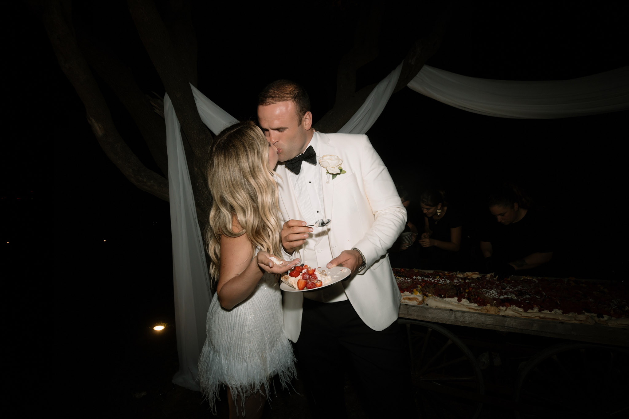 Bride in feathered mini dress and groom in white dinner jacket kiss during strawberry cake cutting