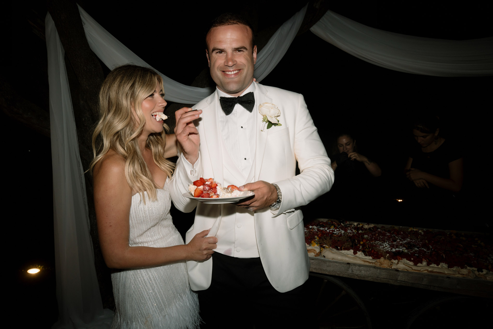 Groom in white dinner jacket feeding bride cake at outdoor evening reception with pavlova dessert table behind