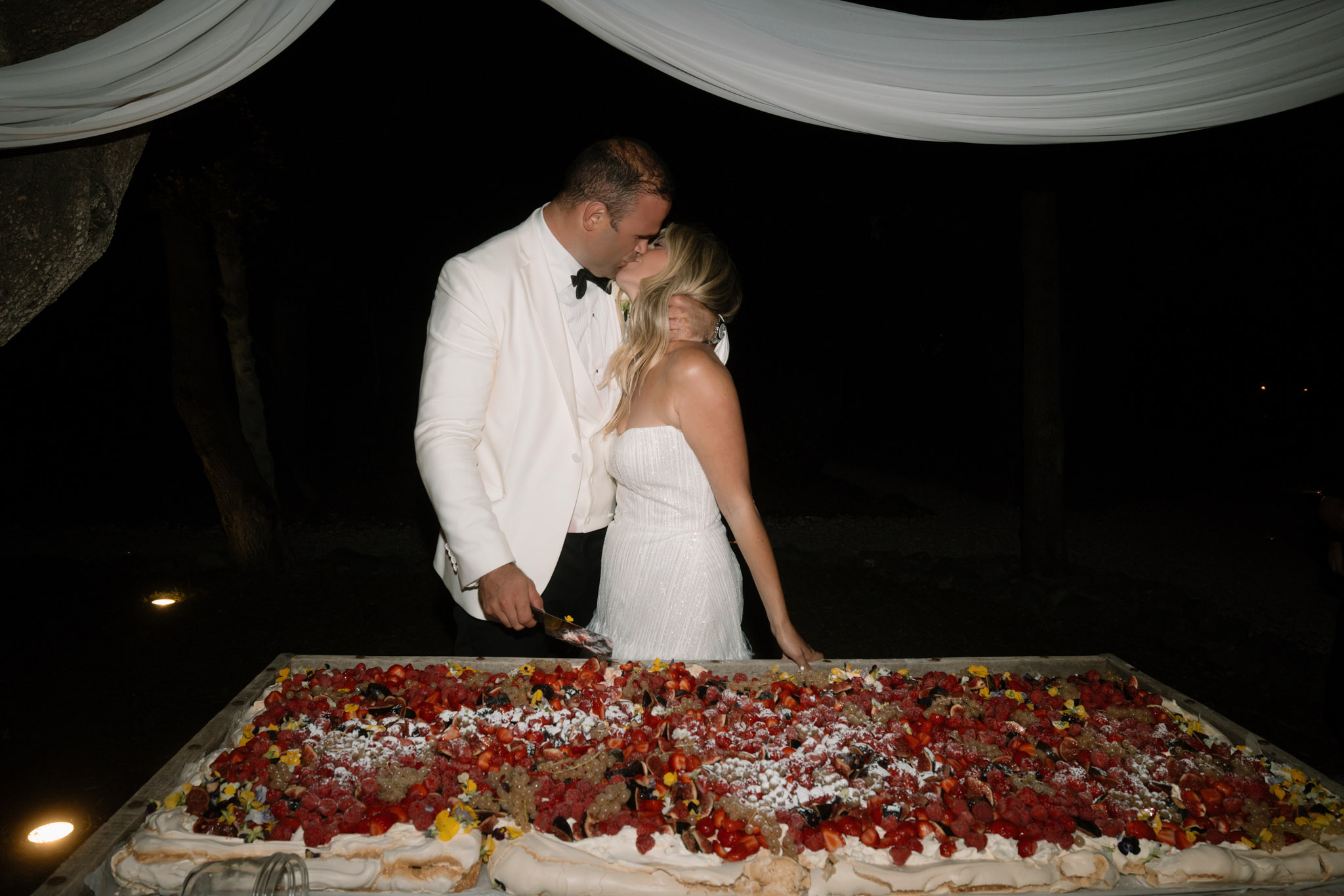 The bride and groom share a kiss during the cake cutting moment at an outdoor evening reception. The groom wears a white dinner jacket with black bow tie and black trousers, while the bride wears a strapless, form-fitting white sequined or beaded gown with long blonde hair worn down. In the foreground is a large rectangular wedding dessert — likely a pavlova or meringue slab cake — topped with whipped cream, strawberries, raspberries, figs, and scattered yellow and purple edible flowers dusted with powdered sugar. The background is nearly black, with white fabric draping visible overhead and small ground-level uplights creating warm spots of light at the base of the frame. The setting appears to be an outdoor terrace or garden area at night. The shot is taken from a medium distance, slightly below the cake level, placing the couple centrally above the dessert.