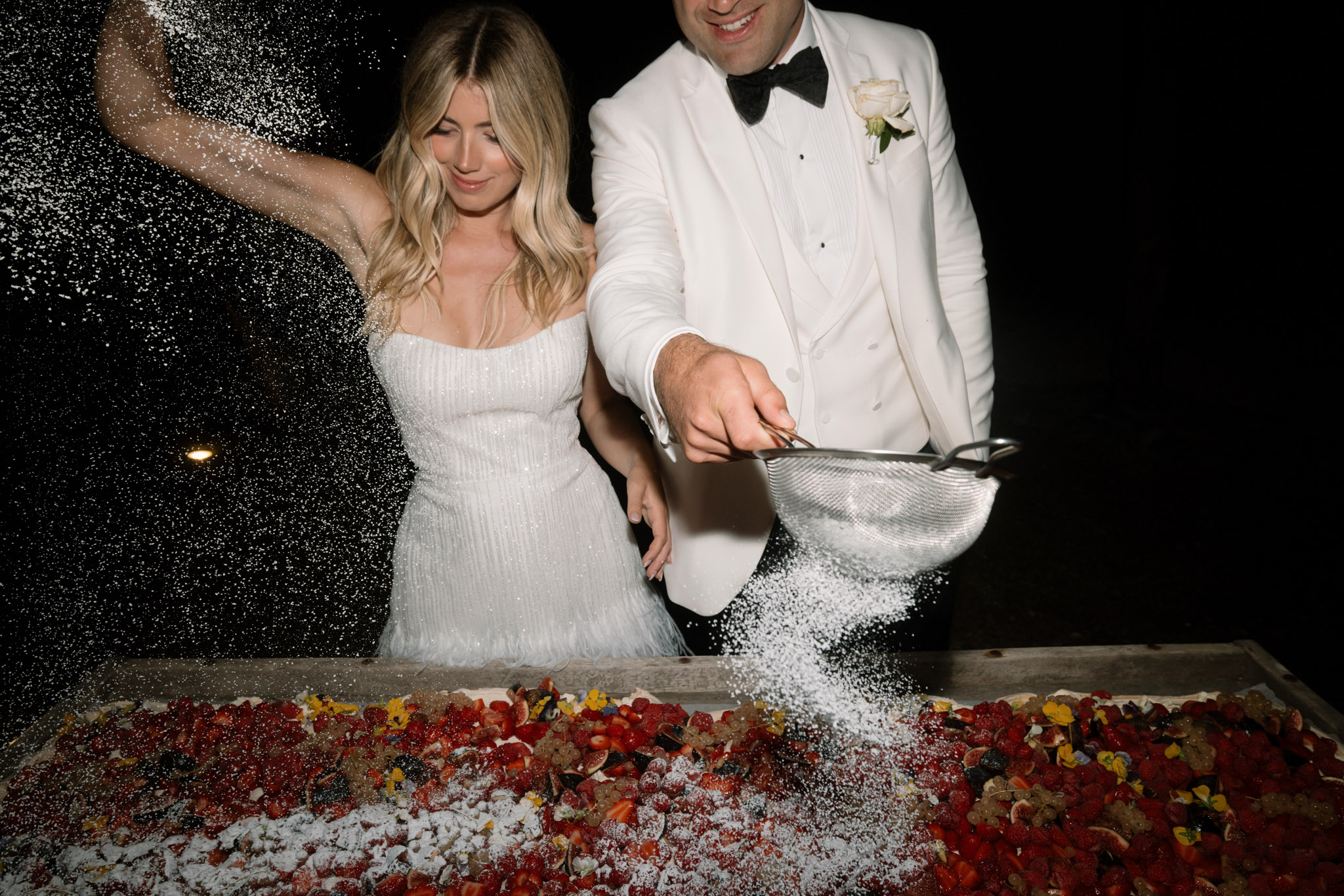 Bride and groom dusting powdered sugar over fresh fruit tart during reception, sugar cascading through air