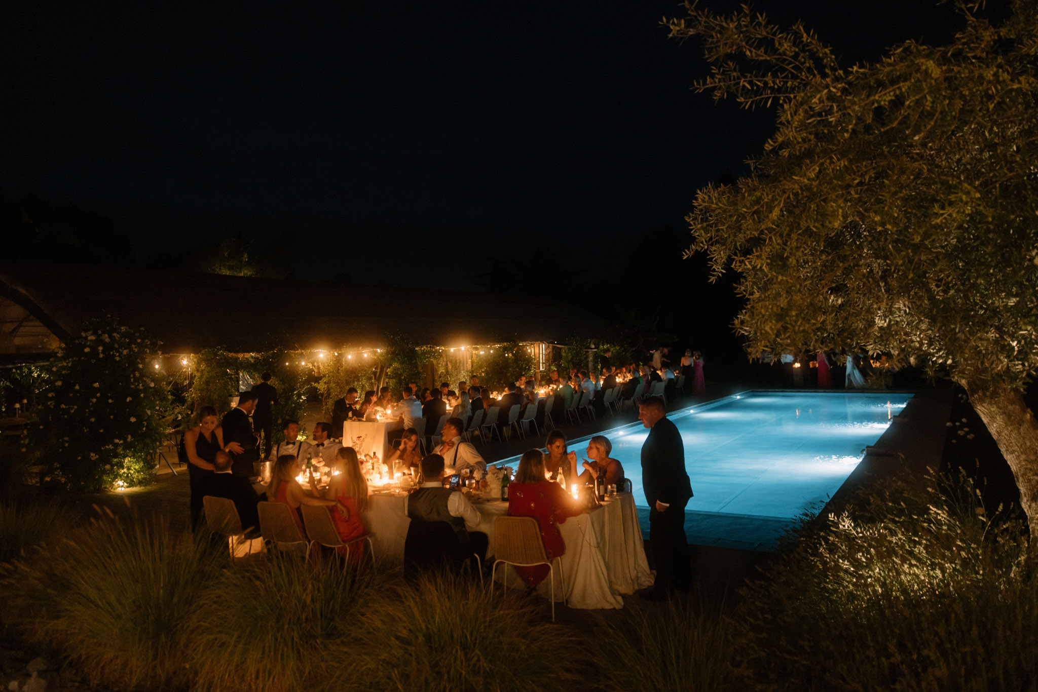 Evening poolside reception sixty guests at candlelit tables with bistro lights lit blue pool and bride in white visible