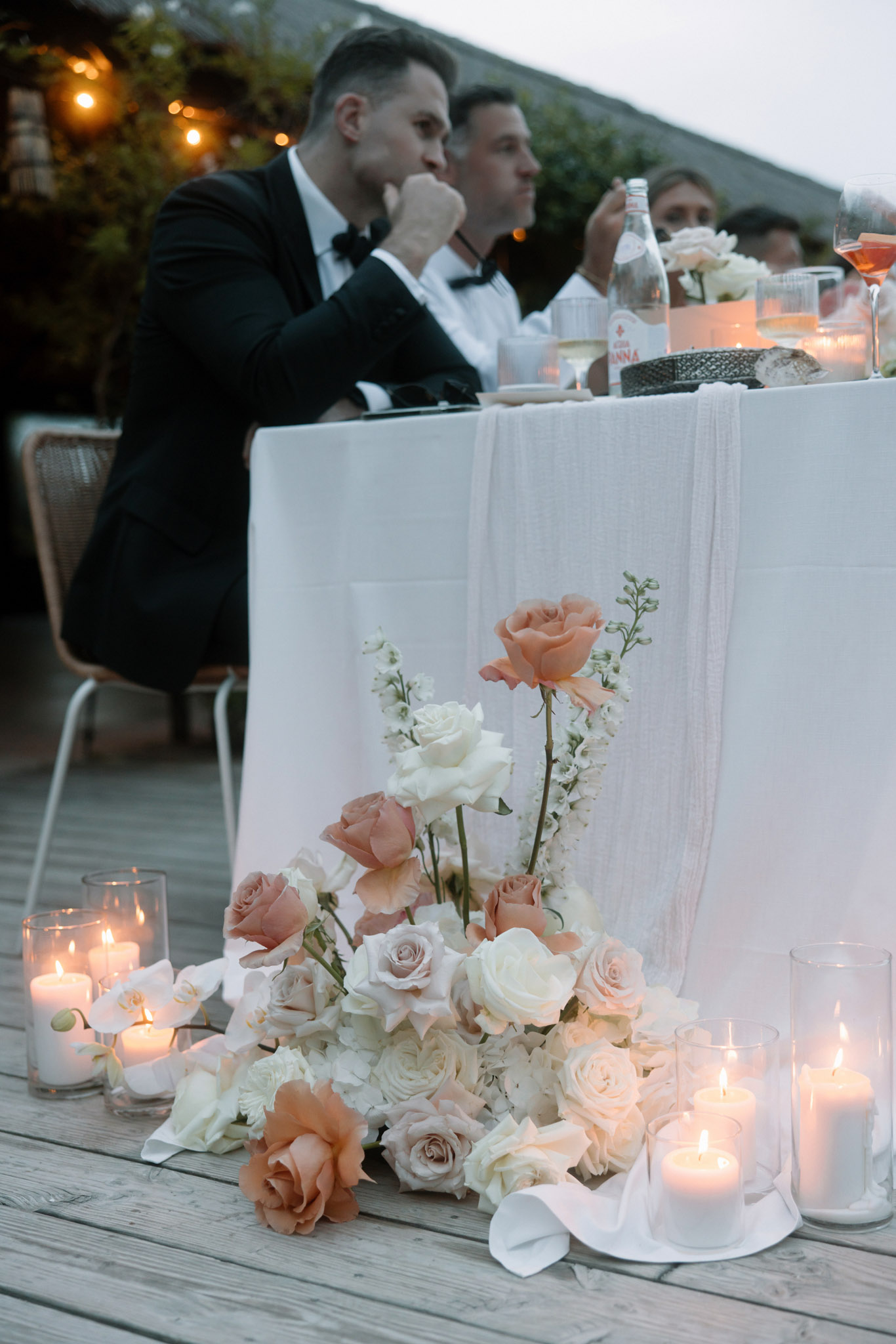 An outdoor evening wedding reception captured in a close-up detail shot focused on floor-level floral and candle decor at the base of a head table. The floral arrangement is a loose, cascading ground-level design featuring peach roses, dusty mauve roses, ivory roses, white stock flowers, and white orchid blooms, spilling across a wooden deck with white ribbon or fabric weaving through. Multiple pillar candles in clear glass hurricane vases are lit on both sides of the arrangement, creating warm candlelight at dusk. The head table is dressed in a white linen with a draped white gauze runner, set with Acqua Panna water bottles, wine glasses, and a rose-tinted cocktail glass. In the soft-focus background, two male guests in black tuxedos with bow ties are seated at the table alongside other guests, with warm string lights visible further behind. The overall decor palette is ivory, peach, and dusty rose with a modern classic styling approach.