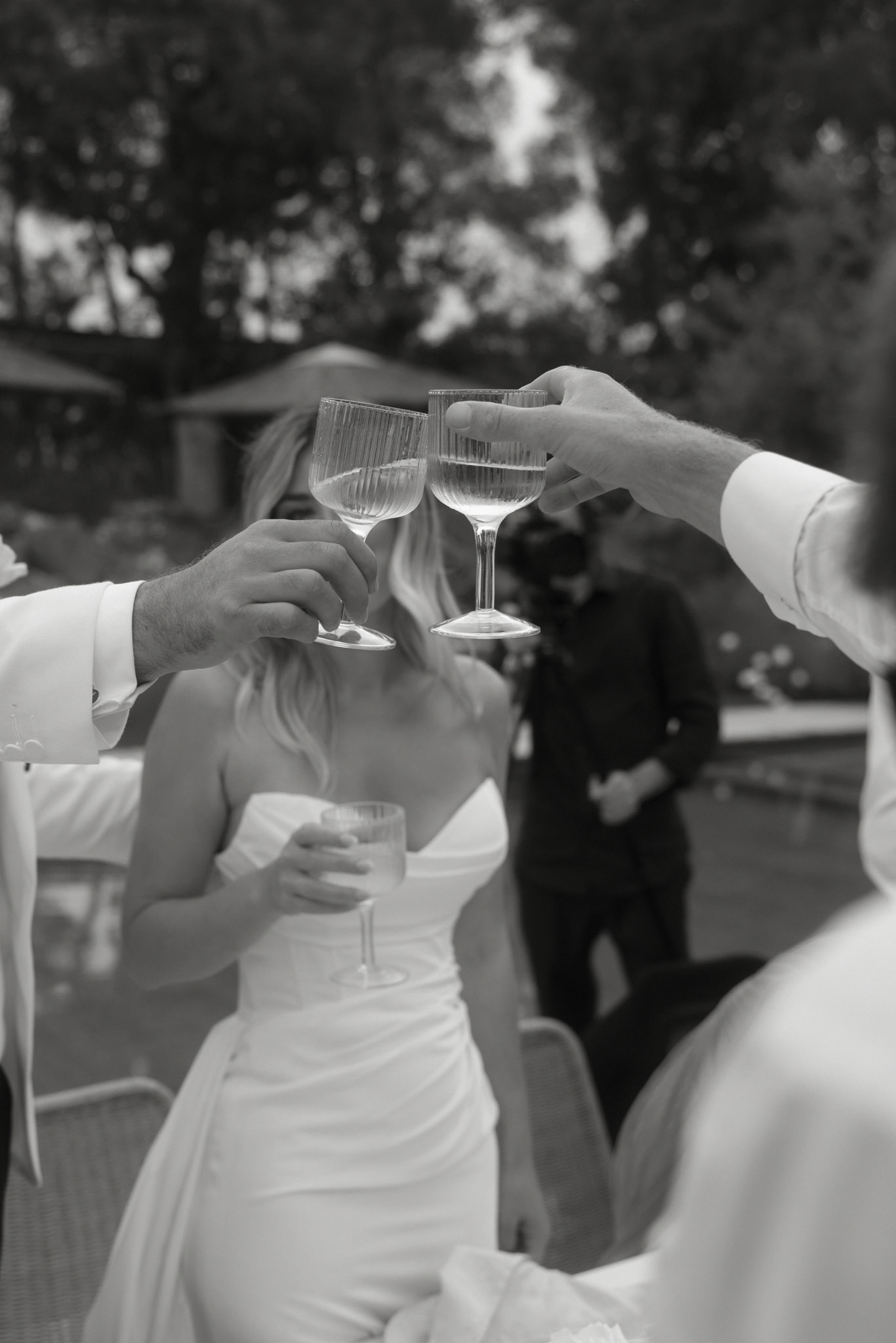 Black-and-white close-up of coupe glasses being clinked during a toast with bride seated between guests