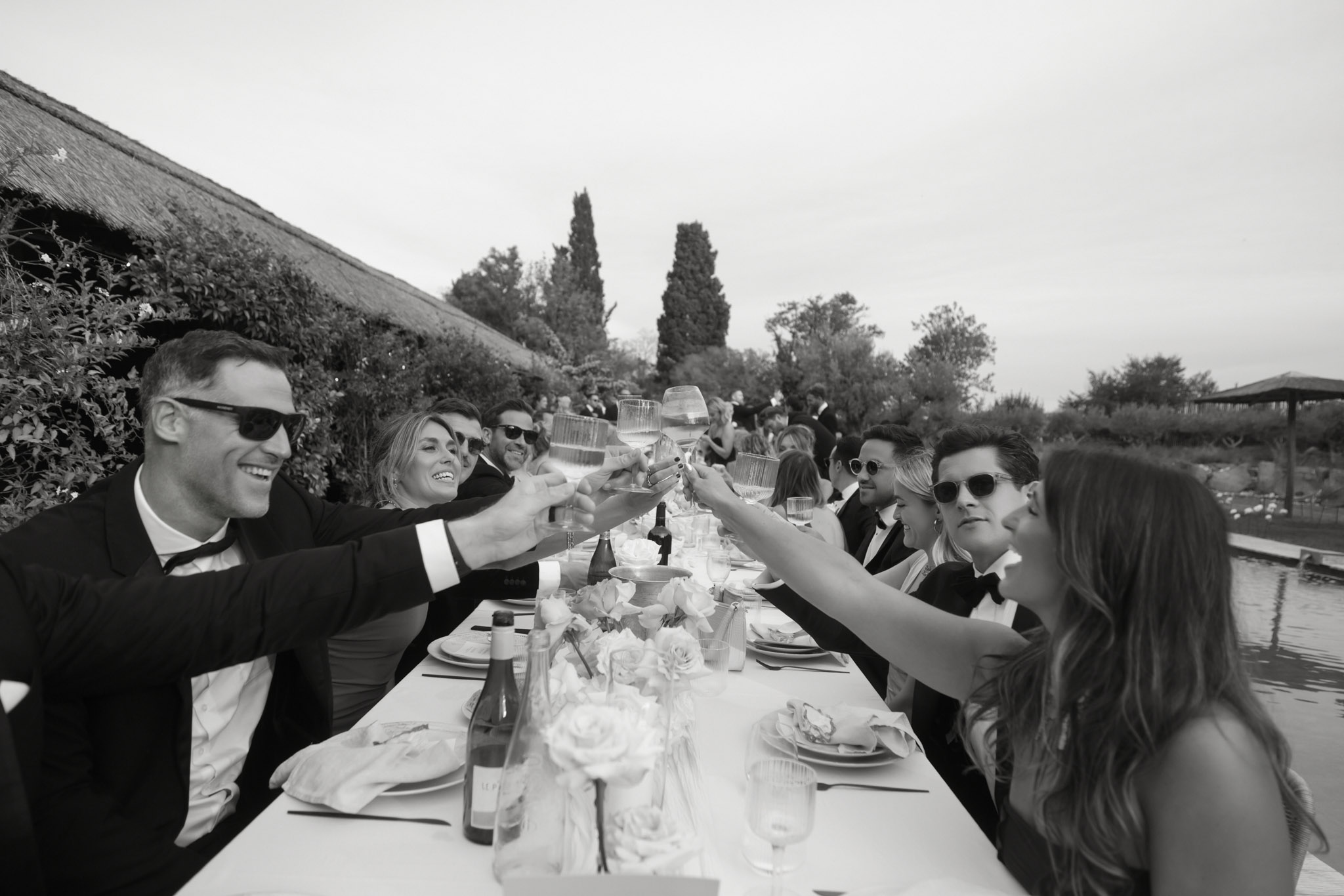 Guests in black tuxedos raising glasses for toast at long outdoor reception table in black and white