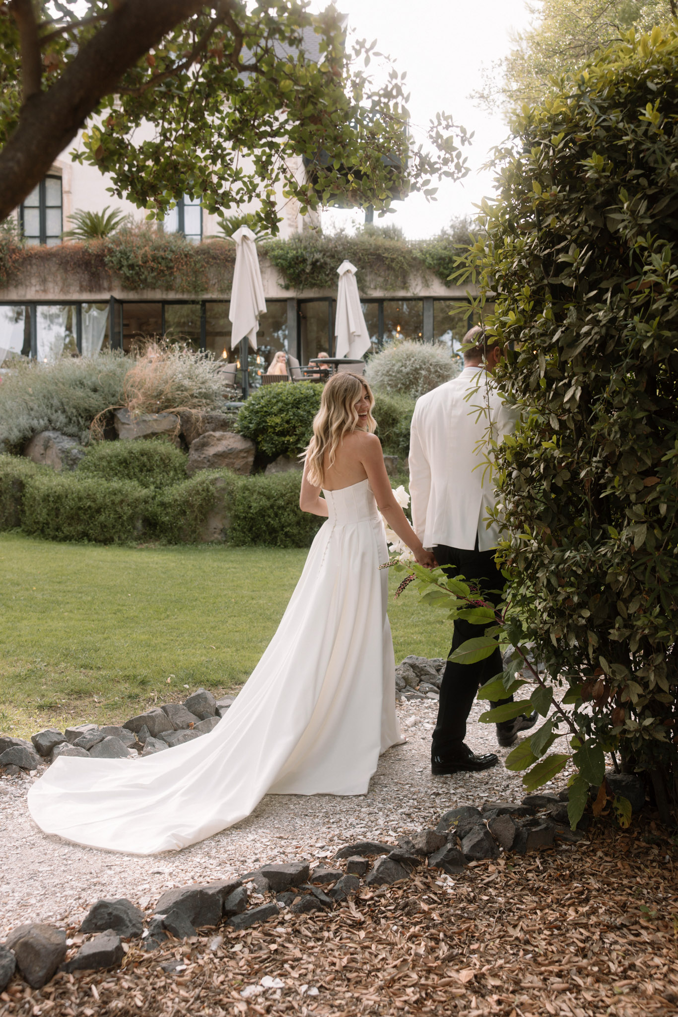Bride in strapless gown and groom in white dinner jacket walking hand-in-hand along garden path