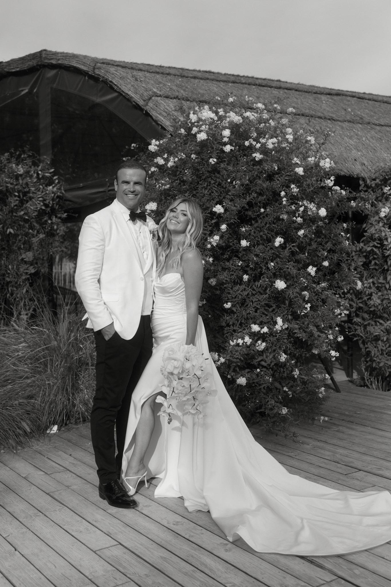 This is a black-and-white couple portrait taken outdoors on a wooden deck. The bride wears a fitted strapless gown with a flowing train and a high slit, carrying a large rounded bouquet with what appear to be oversized blooms, likely hydrangeas or peonies. The groom is dressed in a white dinner jacket, black trousers, and a black bow tie. They stand close together facing the camera, smiling. Behind them is a large flowering climbing shrub with small light-toned blossoms against a structure with a thatched or reed roof. The image has strong contrast with bright highlights on the white garments and deep dark tones in the foliage. The overall styling is modern and sleek. Full-length portrait composition.