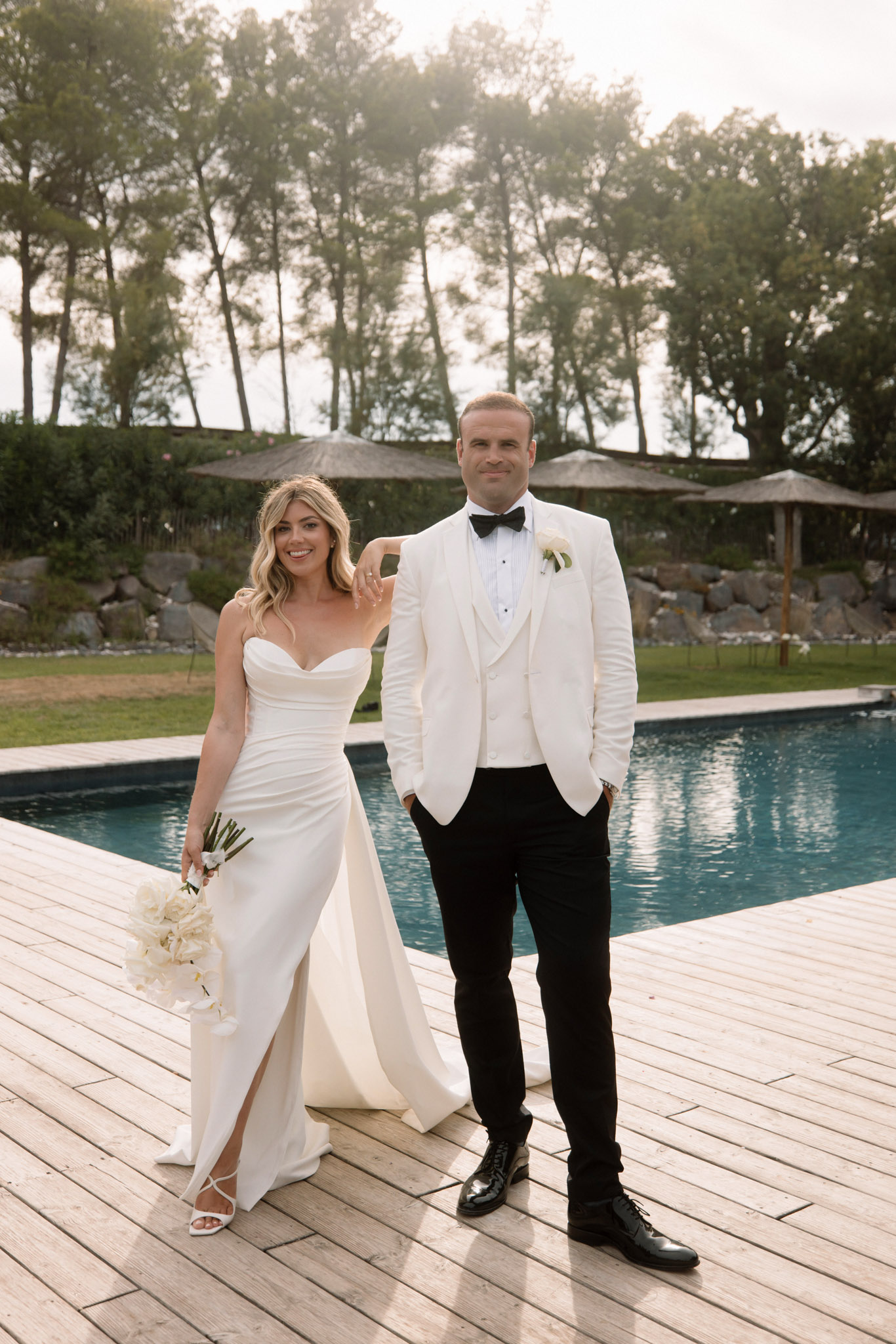 Bride in ivory strapless satin gown and groom in white dinner jacket standing on a pool deck at golden hour