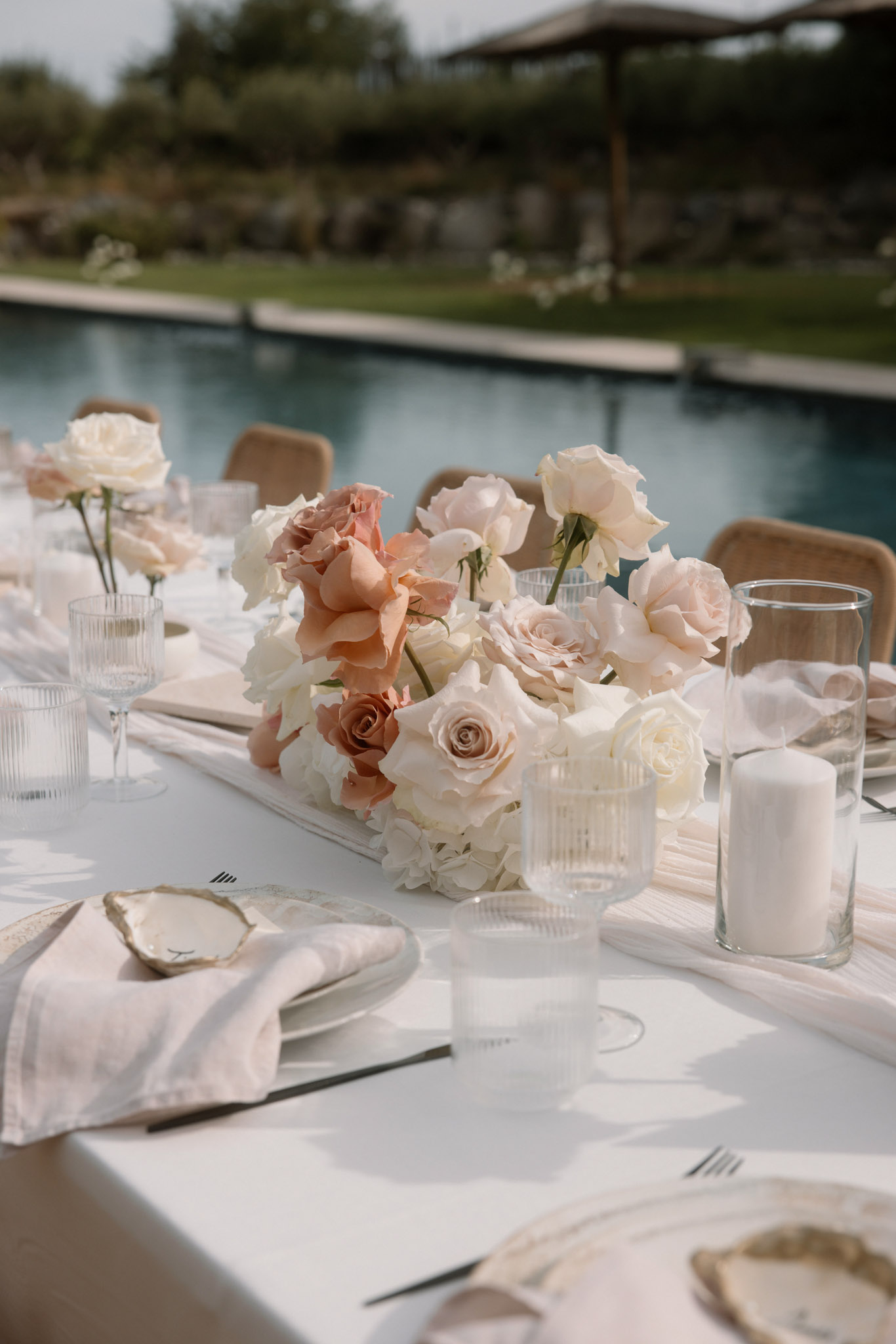 Close-up detail shot of an outdoor wedding reception table set beside a pool. The centerpiece features a low arrangement of blush pink, peach, ivory roses, and white hydrangeas displayed in a ribbed glass vase. The table is dressed with a white linen cloth, white plates, blush linen napkins, gold-edged oyster shell place card holders, ribbed glassware, and white pillar candles in clear cylinder vases. Single ivory and blush roses in small glass bud vases are also visible along the table runner. The overall decor palette is ivory, blush, and peach with natural gold accents, reflecting a modern minimalist style. Rattan-backed chairs are partially visible in the background, and a rectangular pool is out of focus behind the table.
