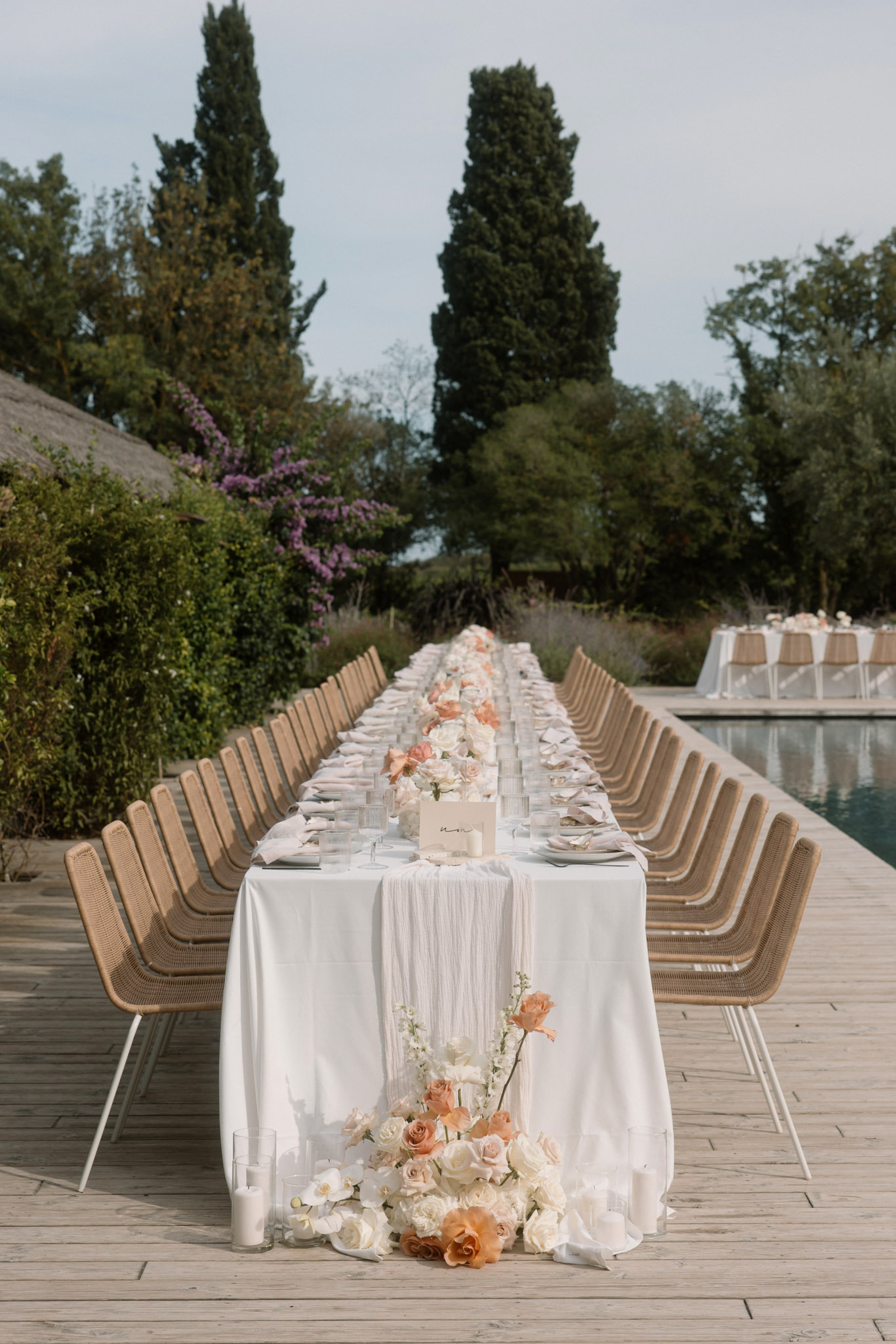 Long feasting table with peach and ivory rose centerpieces on wooden deck beside pool at outdoor reception