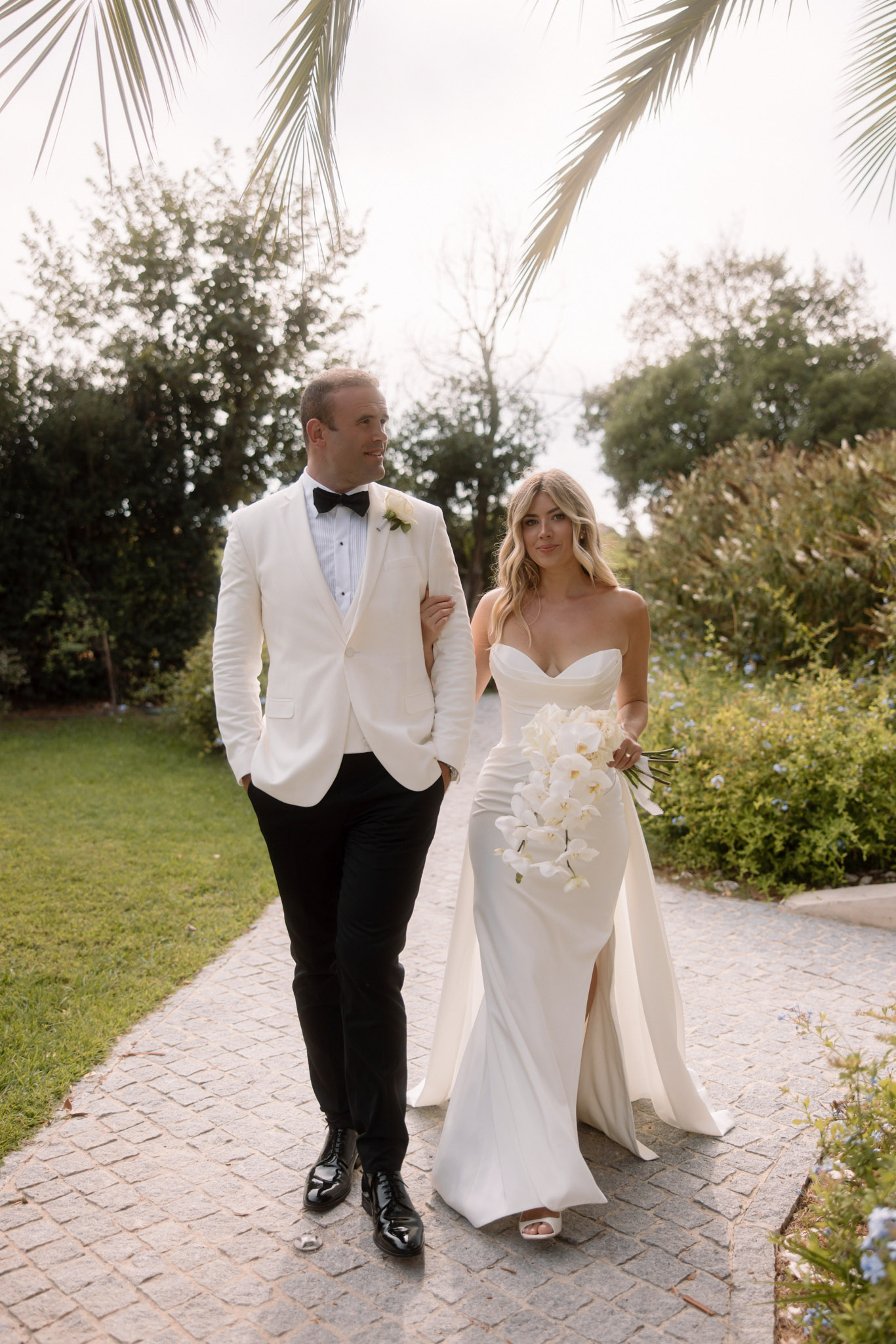 A couple portrait taken outdoors on a cobblestone garden path, with the bride and groom walking arm in arm toward the camera. The groom wears an ivory dinner jacket with black trousers, a black bow tie, and a white floral buttonhole, paired with black patent leather shoes. The bride wears a fitted ivory strapless satin gown with a thigh-high slit and an attached train, carrying a cascading bouquet of white phalaenopsis orchids. The styling is modern and clean with a monochromatic ivory and black palette. Palm fronds frame the upper portion of the image in a medium full-length portrait composition shot in natural daylight.