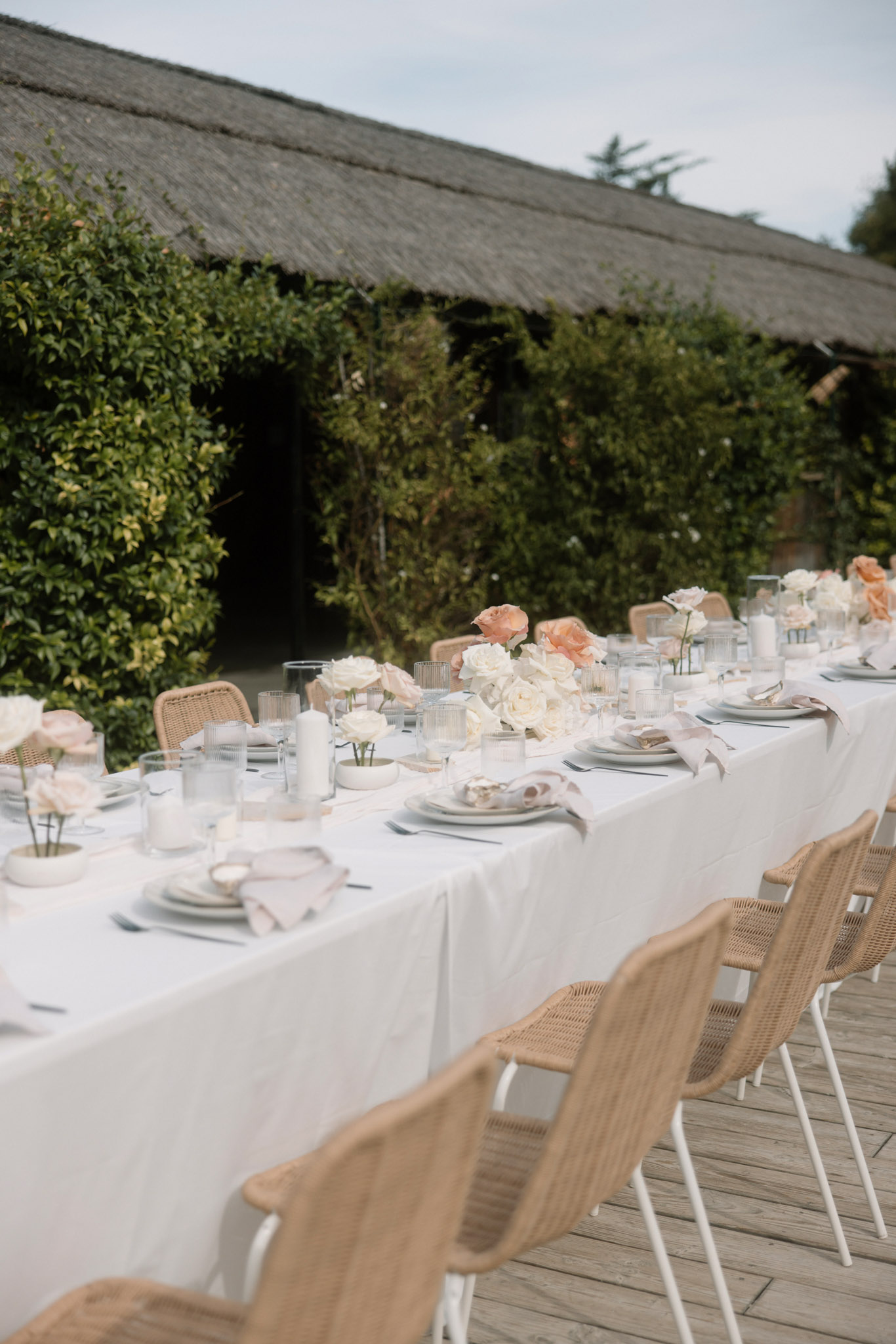 Long outdoor reception table with ivory and peach roses, rattan chairs, blush napkins, and white taper candles