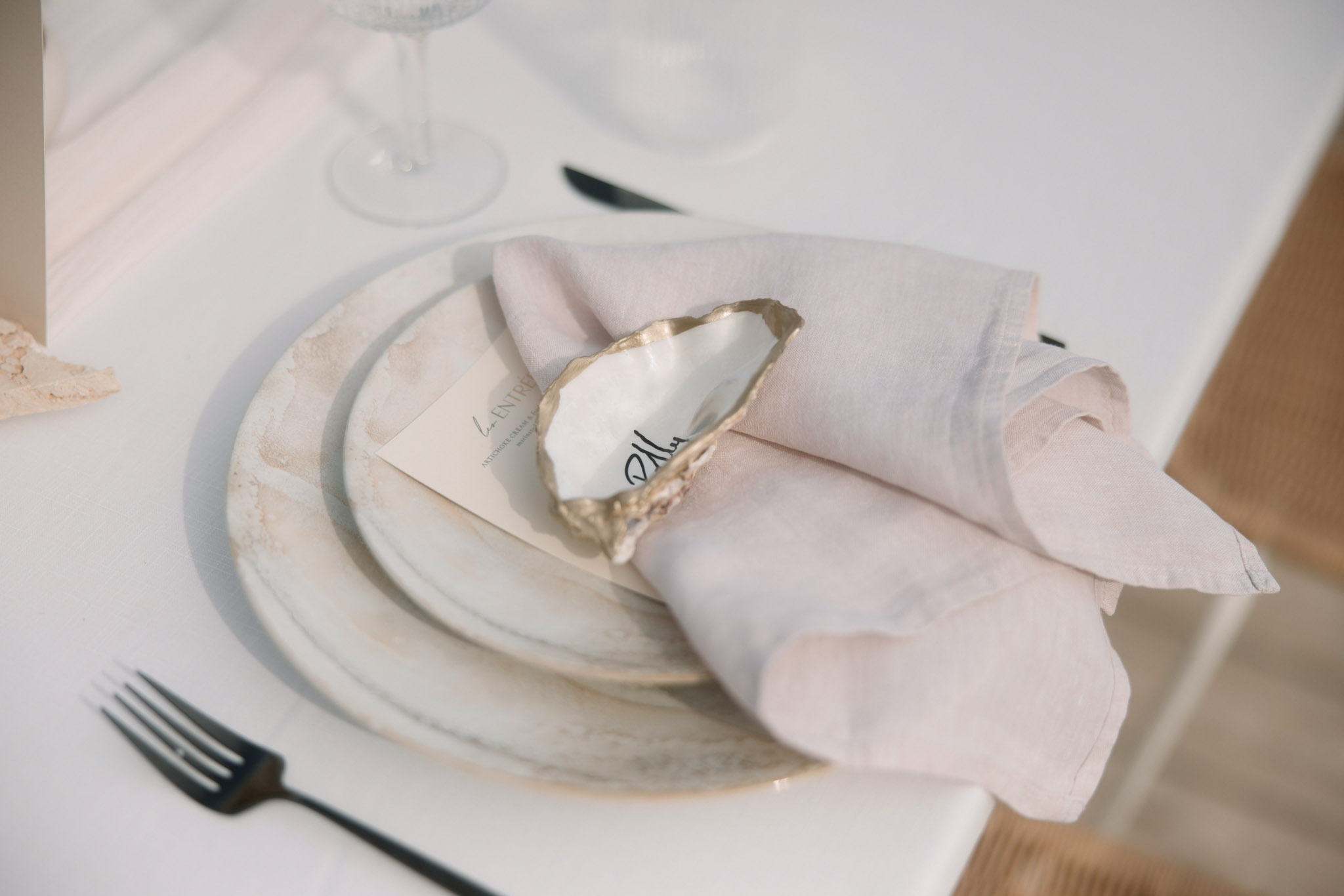 Wedding place setting with stacked ceramic plates, blush napkin, gold-edged oyster shell place card, and black cutlery