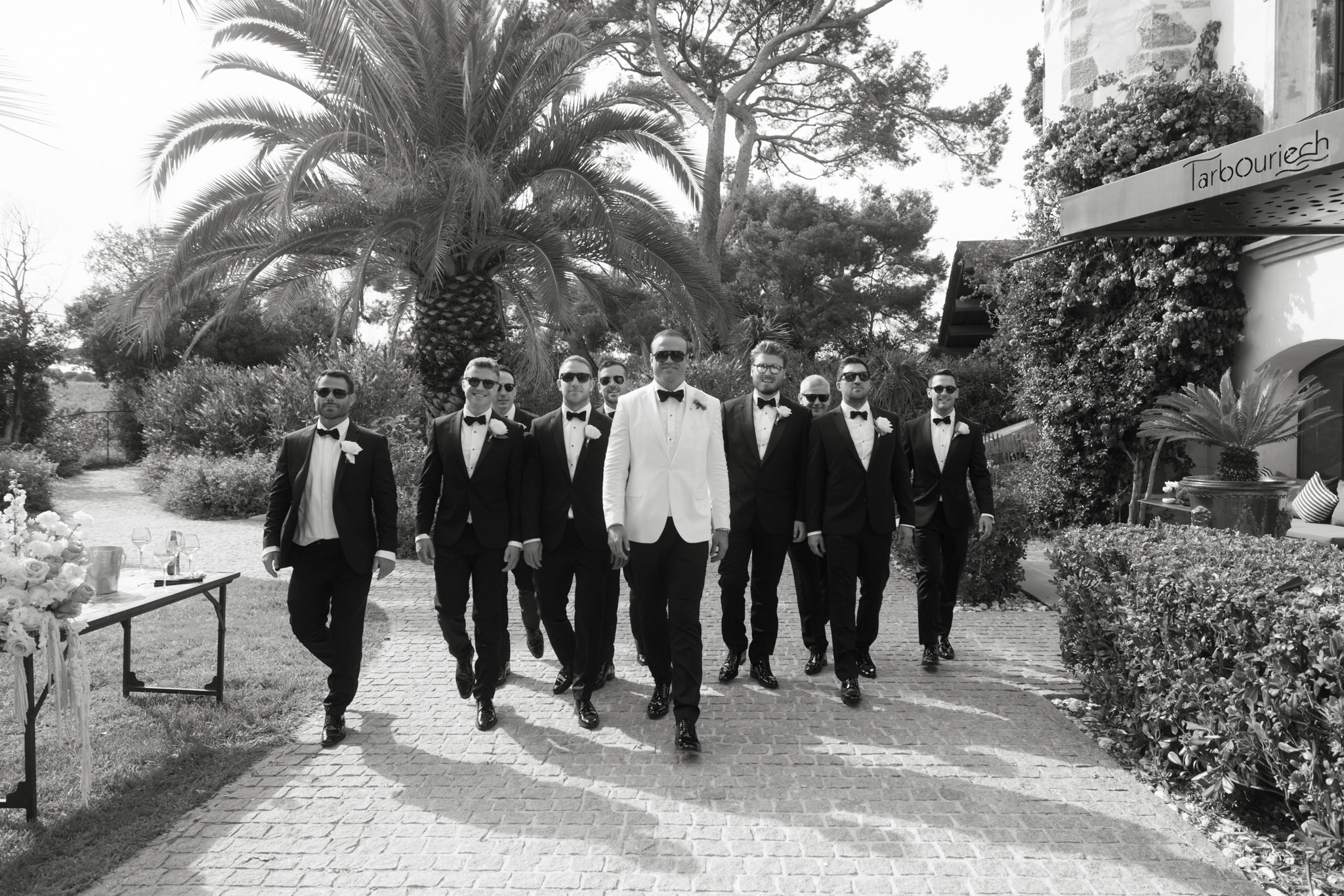Black and white group portrait of groom in white jacket and nine groomsmen in black tuxedos walking together
