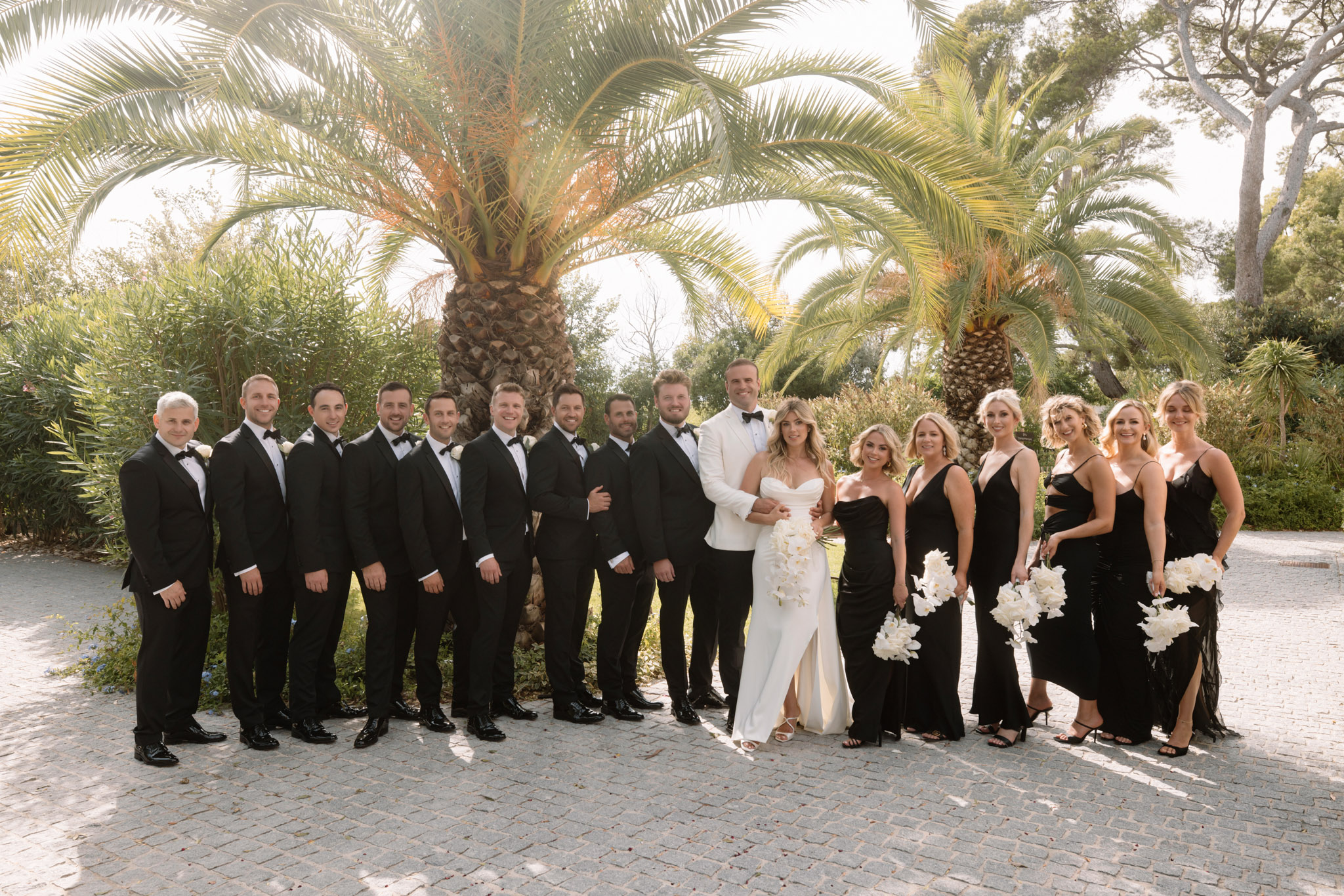 Full bridal party lined up on cobblestone path with bride in white gown and bridesmaids in black dresses