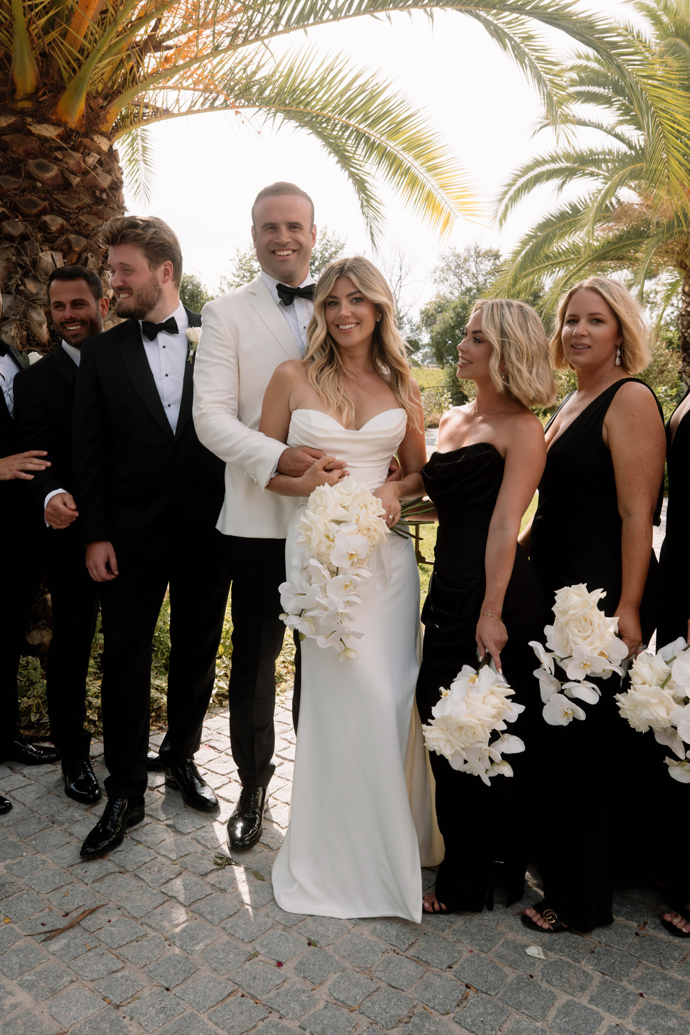 A bridal party group portrait taken outdoors on a cobblestone surface beneath large palm trees. The bride wears a strapless ivory satin fitted gown and holds a cascading bouquet of white orchids and ivory roses; the groom stands behind her in a cream/ivory dinner jacket with black bow tie and black trousers. Visible bridesmaids wear floor-length black strapless and one-shoulder gowns and carry matching white orchid and ivory rose bouquets. Groomsmen wear classic black tuxedos with black bow ties. Approximately six to seven bridal party members are visible in this mid-range portrait shot. The color palette is strictly black, white, and ivory throughout, giving the styling a high-contrast, formal look.